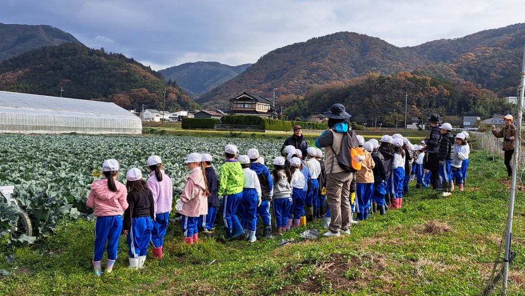 キャベツ畑の前に並び、つちのこクラブメンバーの話を聞いている子どもらの写真