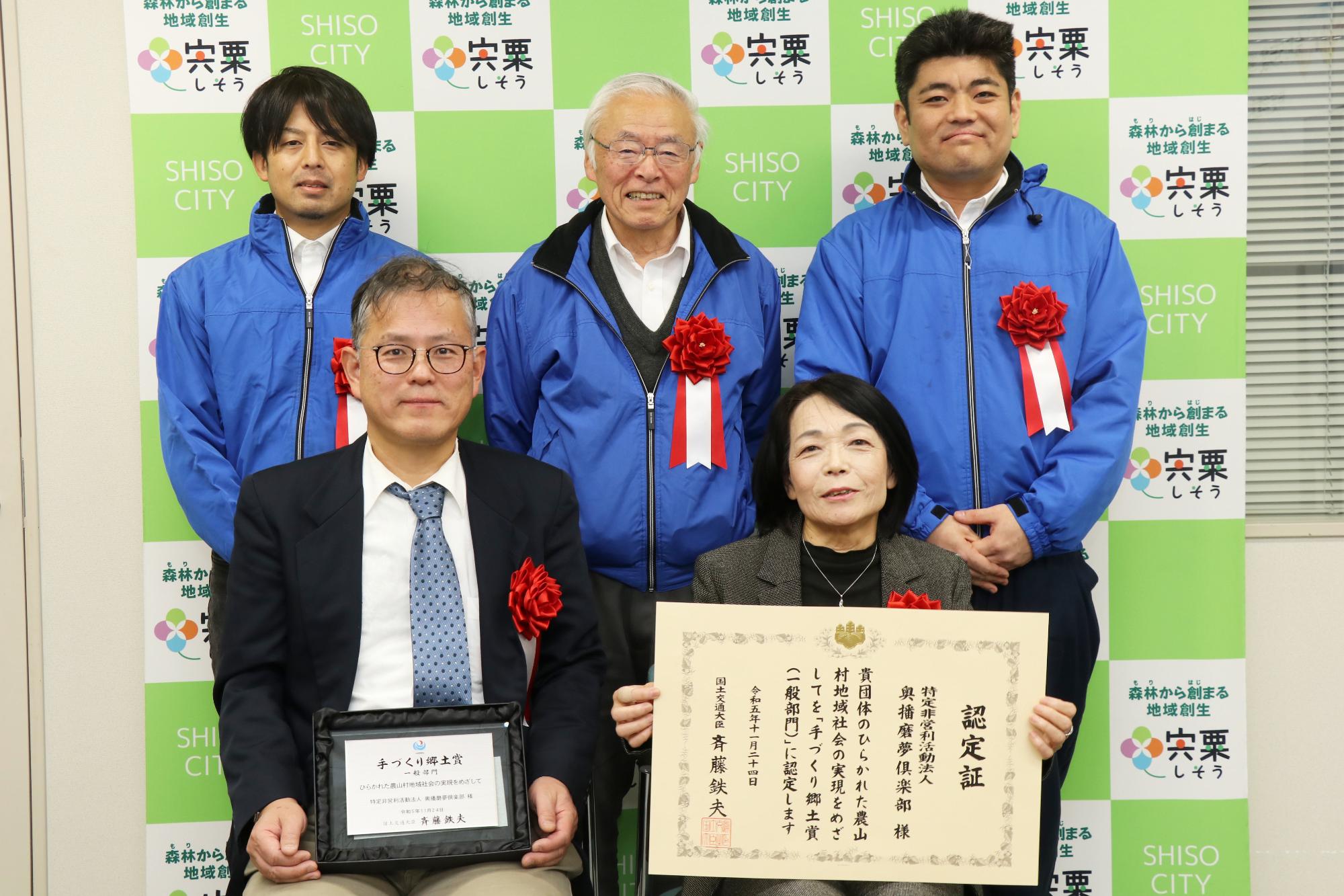 手づくり郷土賞の認定証をかかげた春名さんが前列に座り、その左にひとりが座り、二人の後ろに3人が立って並んでいる写真