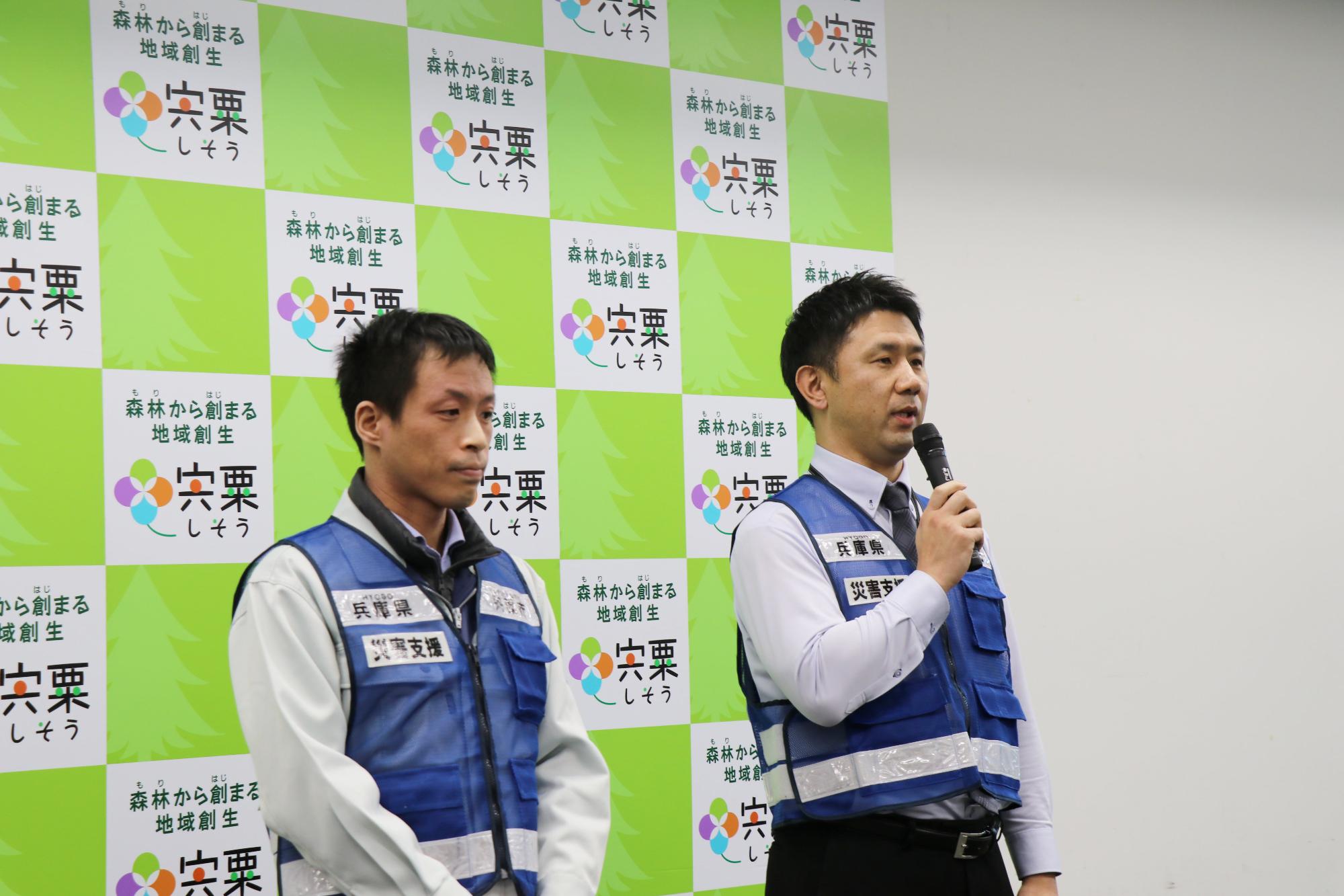 市役所3階の会議室で派遣される職員二人が横に並び、1人がマイクを持って出発表明をしている写真
