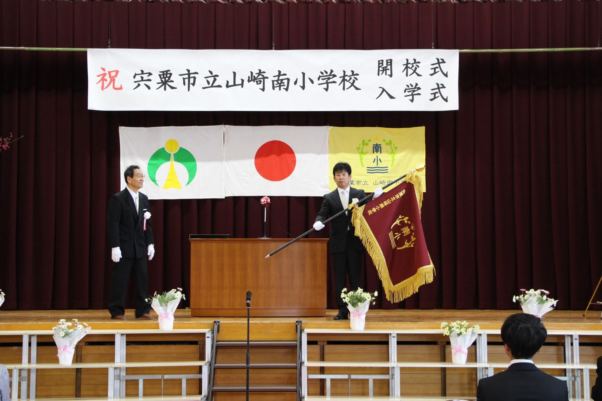山崎南小学校の開校式で校旗を受け取った山本校長と福元市長の写真