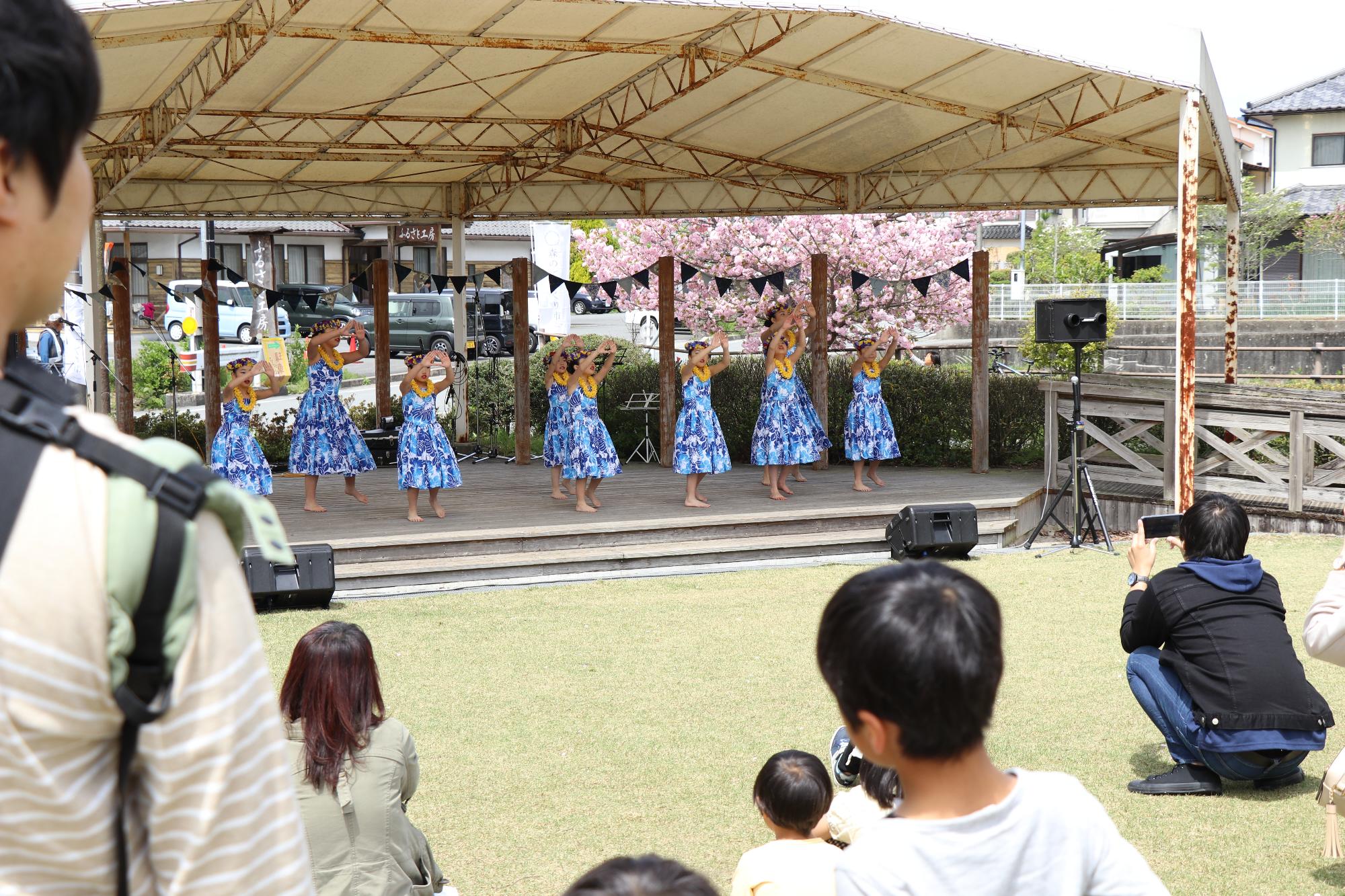ステージ上でフラダンスを披露している水色のスカートを履いた7人の女性と観客の写真