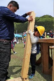 プラスチックの黄色い作業ヘルメットをかぶった男の子が、背丈を超す木の柱を男性の助けを借りつつ台に立てようとしている写真
