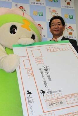 記者発表時にご当地キャラとスーツの男性が「宍粟市に来てね!」ゴム印が押された大きなハガキをカメラに向けている写真