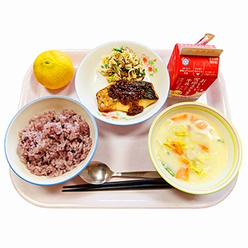 紫黒米のごはん、牛乳、さわらのしょうゆこうじソース、切り干し大根のサラダ、野菜と豆乳のクリームスープ、みかんがお盆の上に並んでいる給食の写真