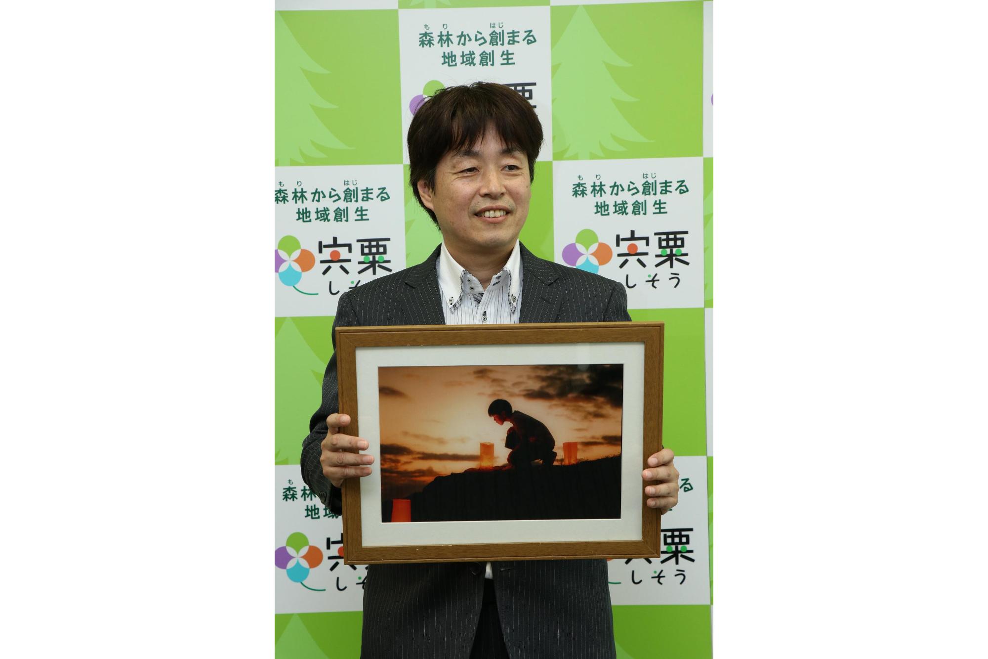 最優秀グランプリを受賞したスーツ姿の小林さんが笑顔で作品を手にしている記念写真