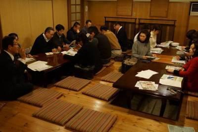 板張りの床と木製の壁の部屋に長い掘りごたつがあり、そこに座って話し合いをしている15人ほどの参加者の写真
