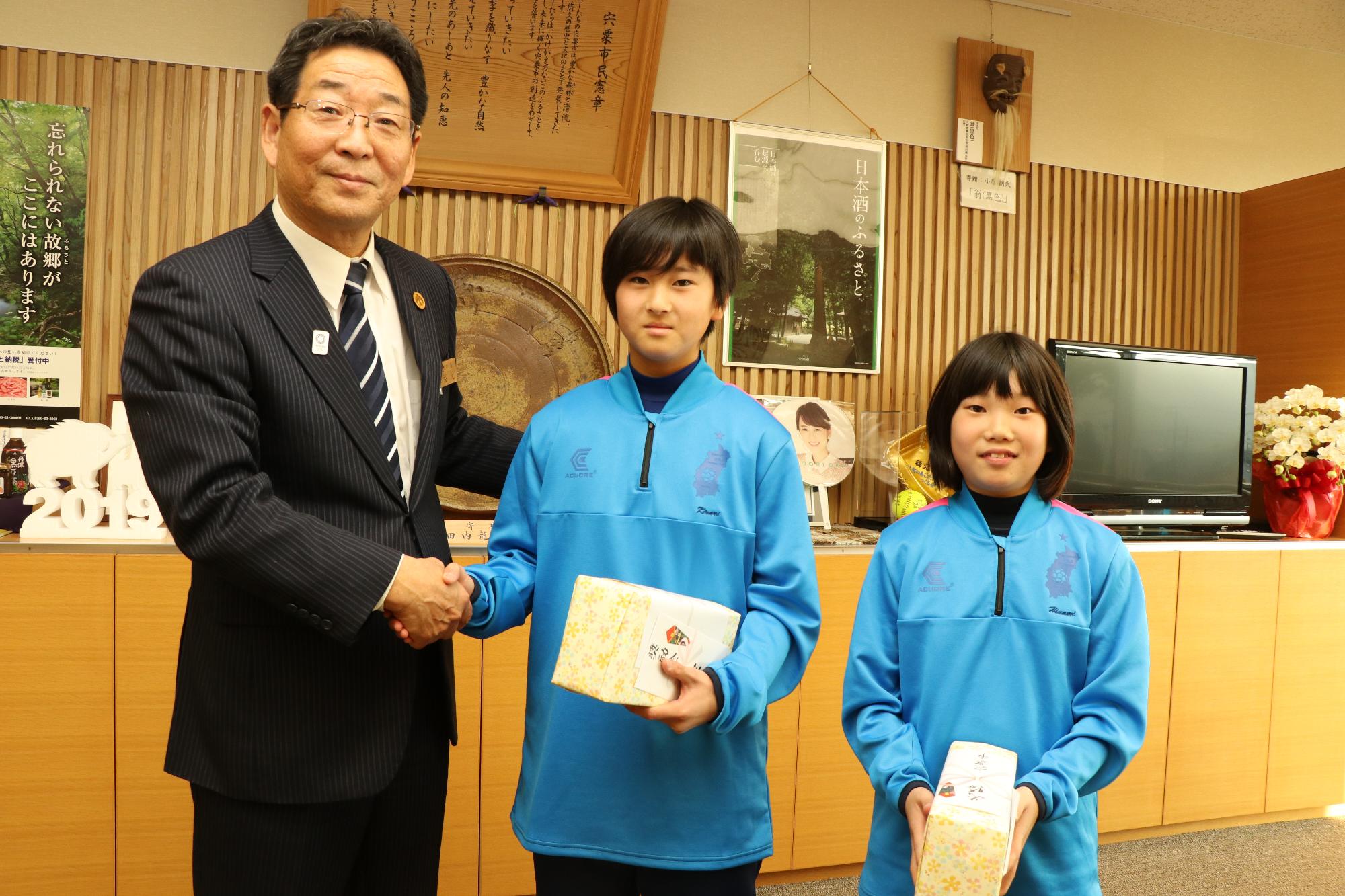 青いナイロン製のジャケットを着た小学生女子サッカーチームの2人と市長の記念写真