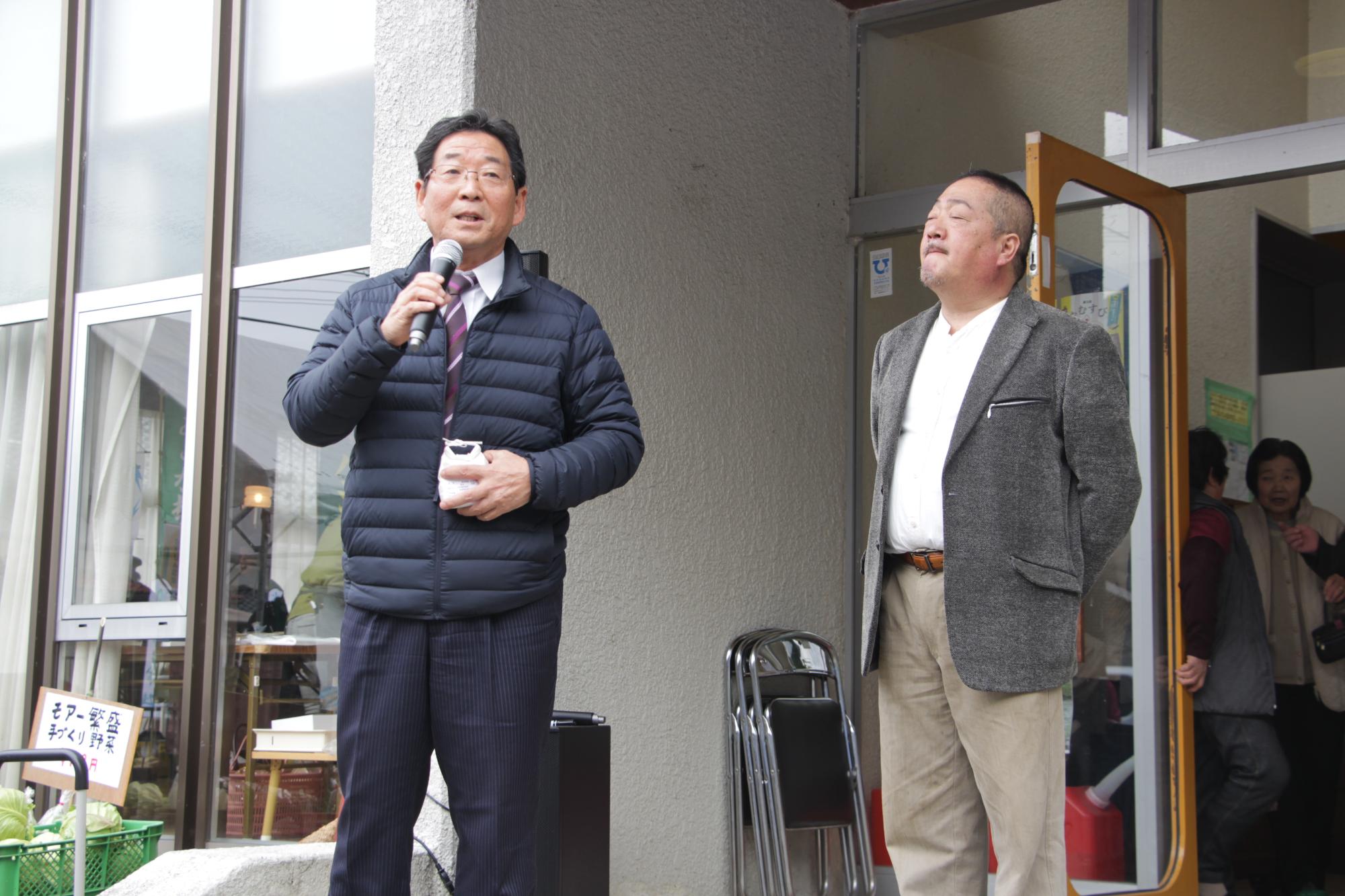 黒いダウンジャケットを着てスピーチをする市長とグレーのジャケットを着た男性の写真