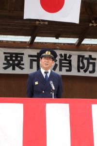 警察の帽子のようなものをかぶり後ろに手を組んだ姿勢の市長が野外に設置された演壇からスピーチをしている写真