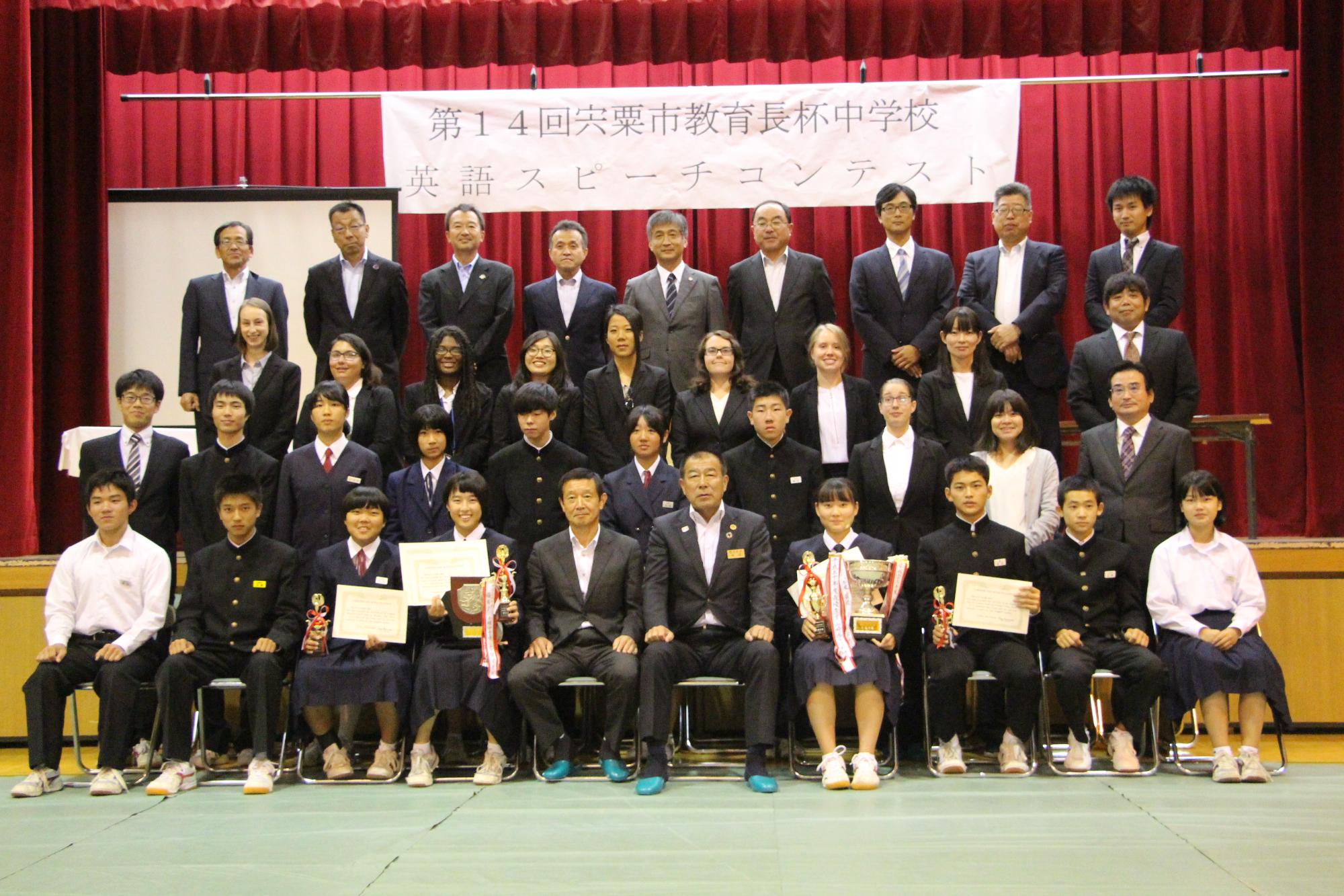 令和元年英語スピーチコンテストに出場した生徒と指導した先生らがひな壇で記念撮影している写真