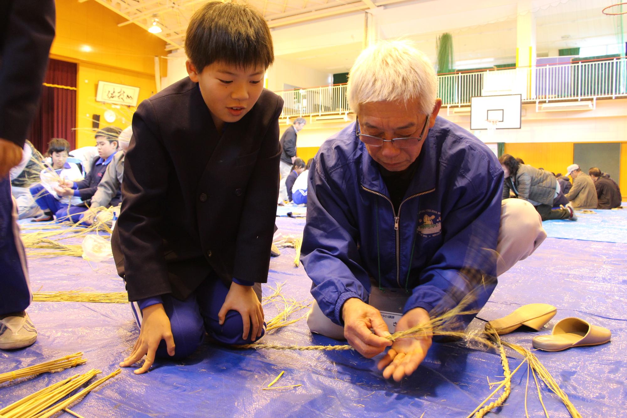 おじいなさんからしめ縄の作り方を教えてもらっている小学生の男の子の写真