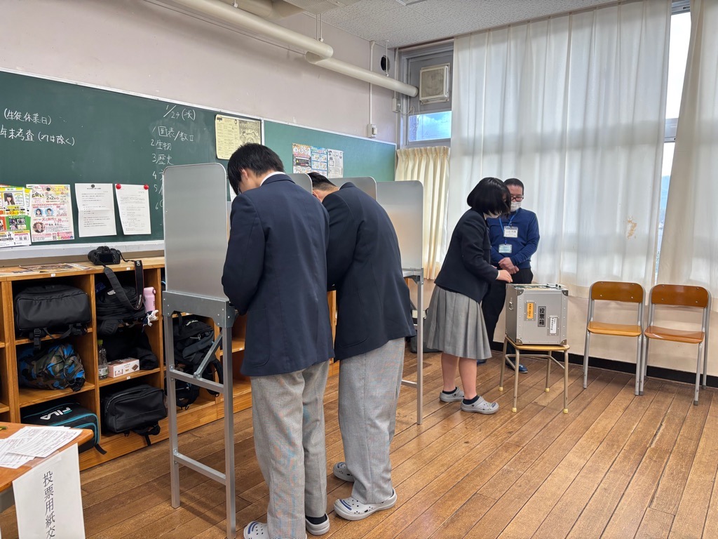 生徒たちが実際の投票で使用される記載台や投票箱を使用して模擬投票をしている様子の写真
