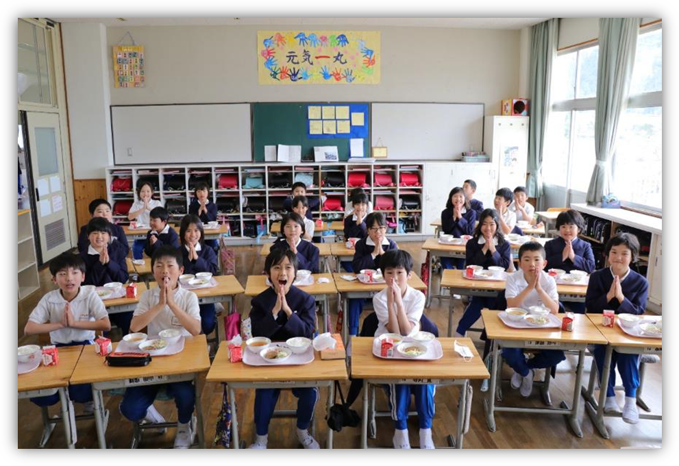 学校の教室で給食を今から児童らが手をあわせて「いただきます」をしている写真