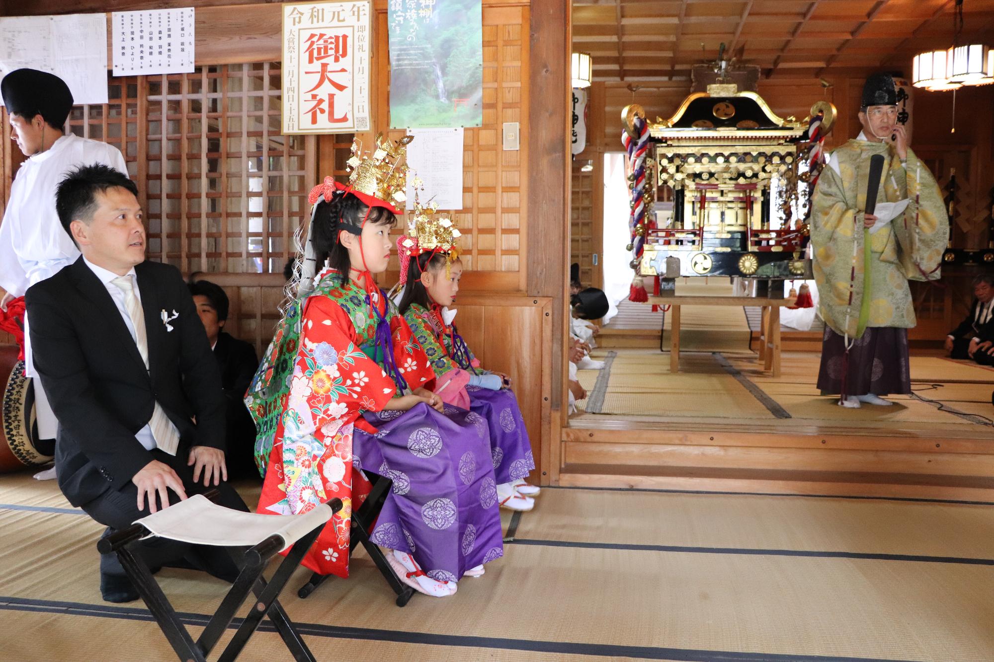 社殿で祭りの始まりを待っている少女神子とその父親の写真