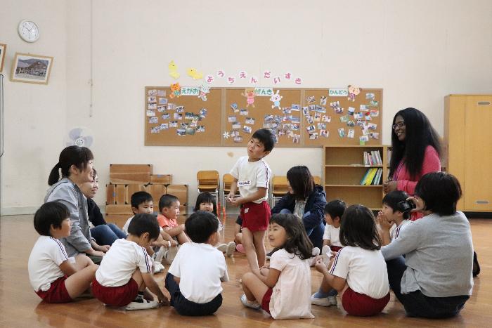円状に内側に座っている子ども達の中央に一人がたつゲームをしながら英語を学ぶ園児達の写真