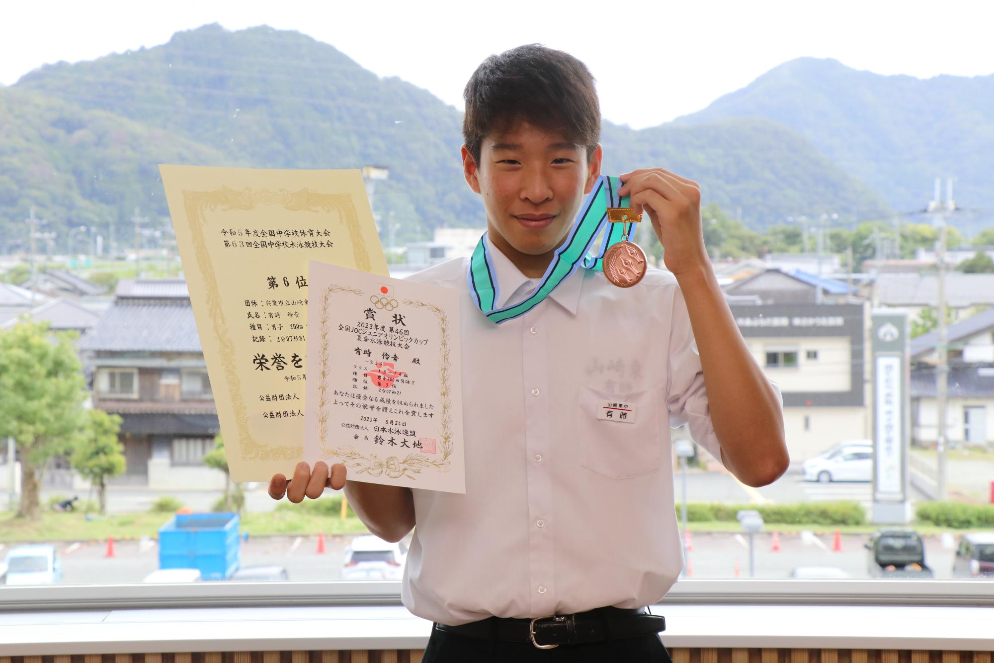 山崎東中学校2年の有時伶音さんが宍粟市役所でメダルを首にかけ、賞状2枚を手に笑顔を見せている写真