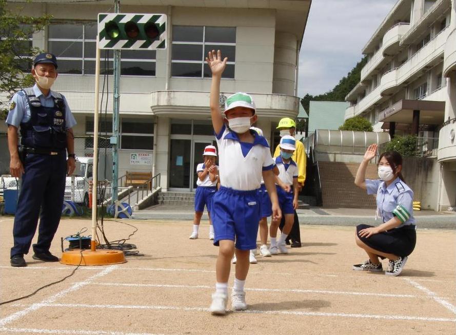 神野小学校の交通安全教室で手をあげて校庭に作られた横断歩道を渡る児童とそれを見守る警察官と指導員らの写真