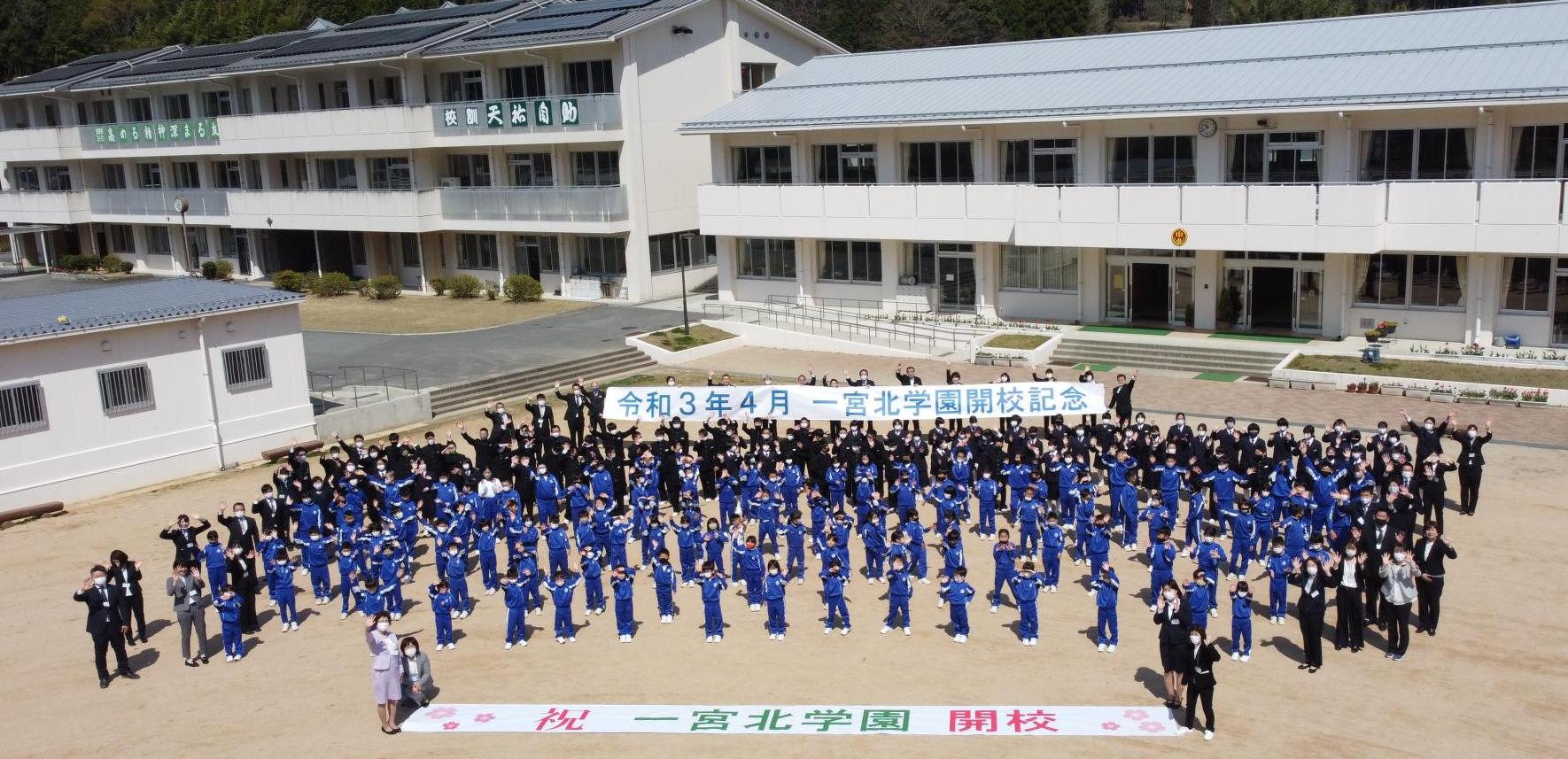 一宮北学園の校舎の前に全校生徒らと教員が並び、一宮北学園開港記念の横断幕を地面においているところを上空から撮影した写真