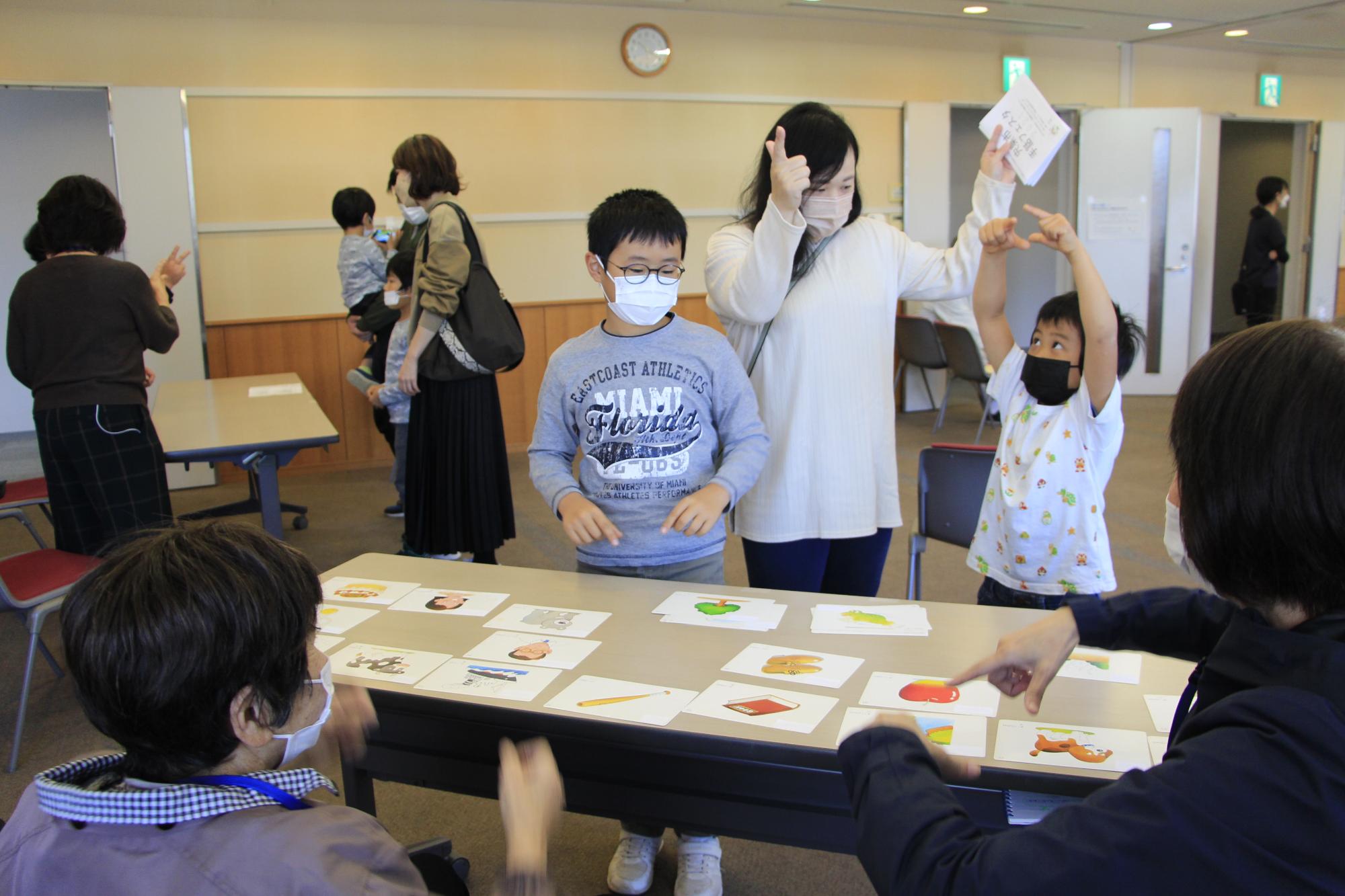 宍粟市手話フェスタでカードに描かれたイラストの手話を講師の女性に教えてもらい指を上に上げている男の子の二人兄弟とお母さんの写真