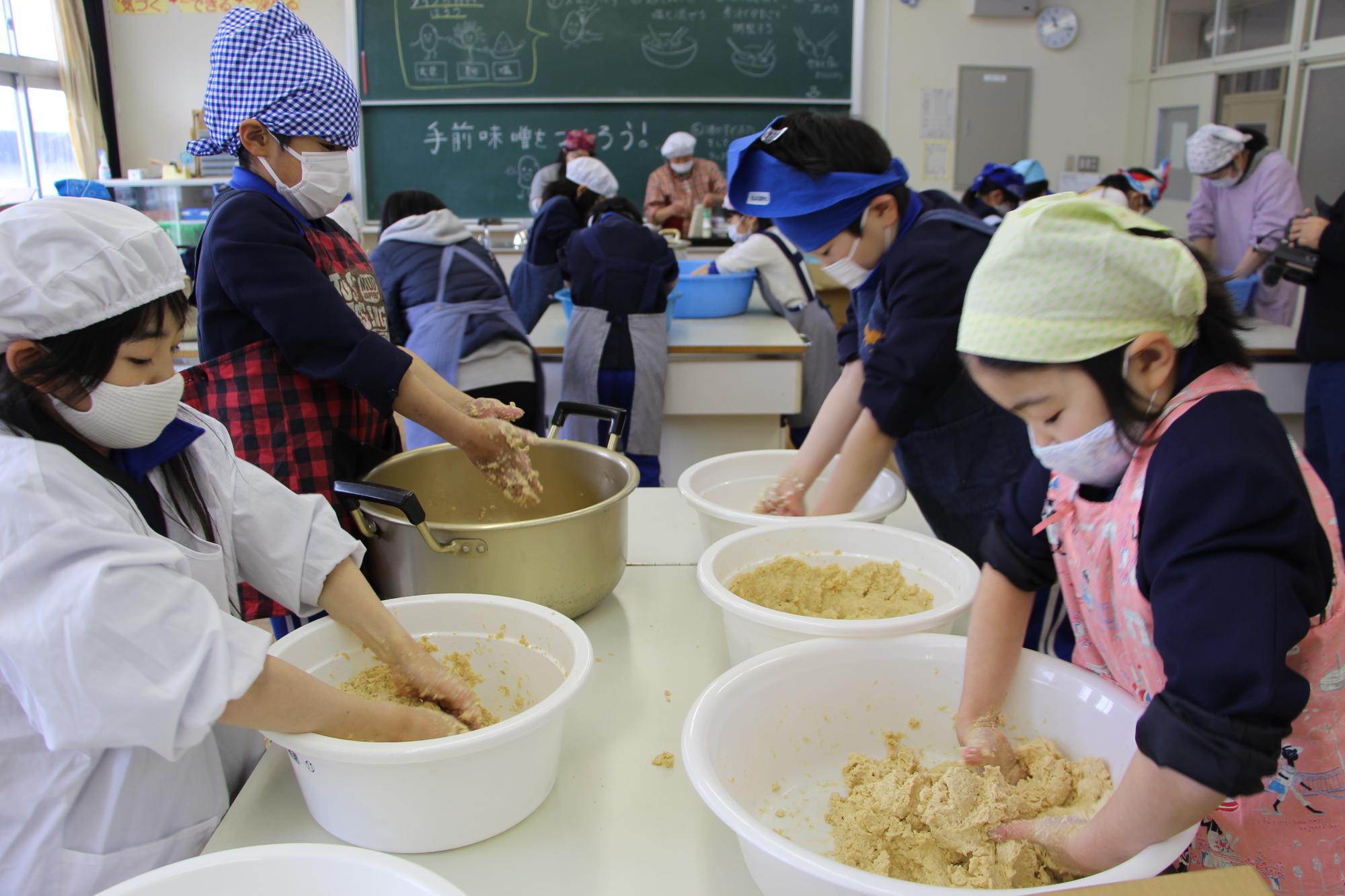 神野小学校の調理室で大豆を手でつぶしながらみその仕込み作業をする子どもたちの写真