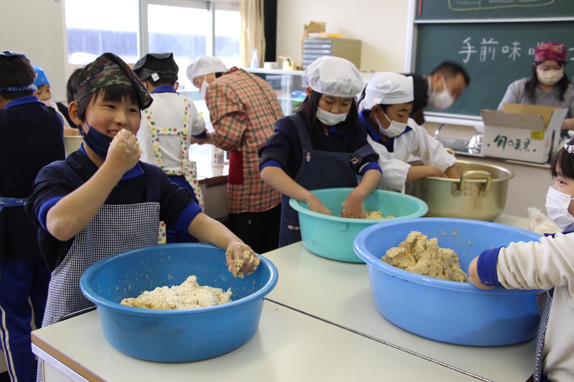 神野小学校の調理室で大豆をつぶしながらみその仕込み作業をする子どもとつぶした大豆の味見をする子どもの写真