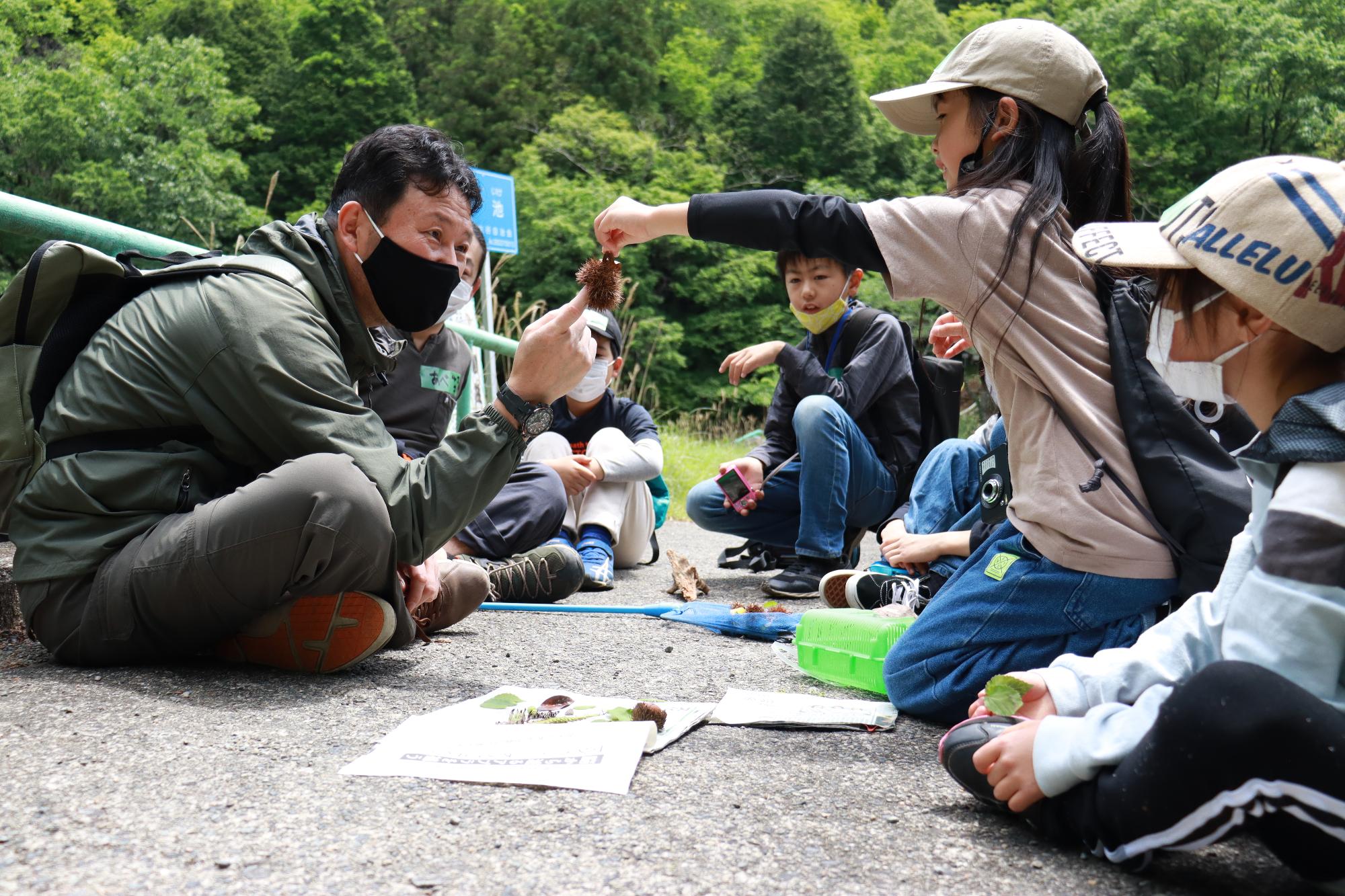 子どもたちが見つけた木の実や葉っぱを地面に並べ、子どもの一人がみんなの前で栗のイガをもって大人に特徴を説明している写真