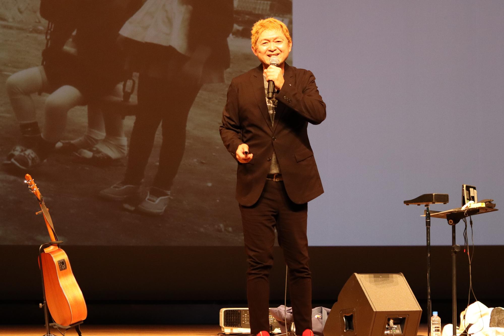 若者フォーラムのステージ上であいさつするシンガーソングライター松本隆博さんの写真