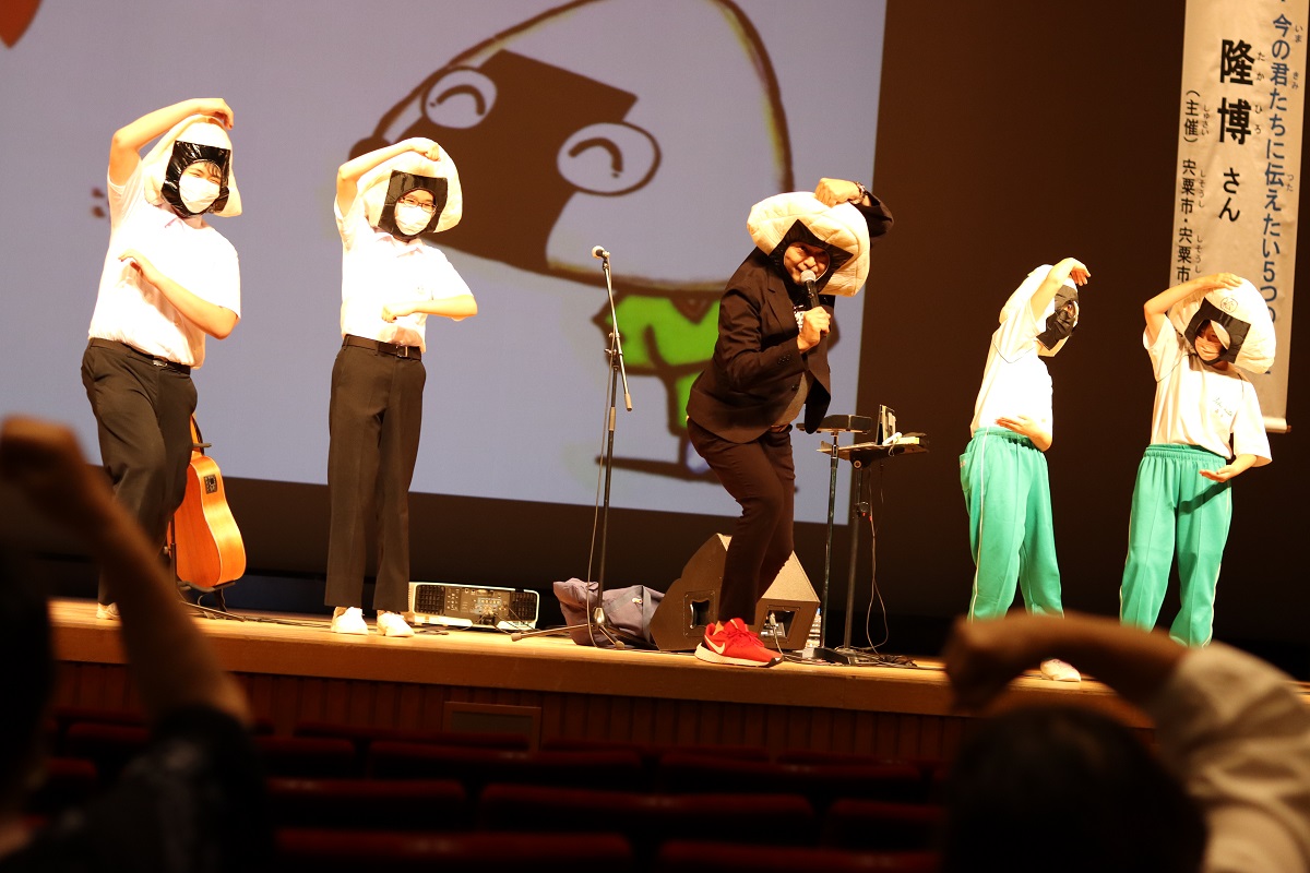 松本隆博さんと4人の中学生がおにぎりのかぶり物を頭につけ、日本人の主食であるお米を食べることの大切さを歌とダンスで表現している写真