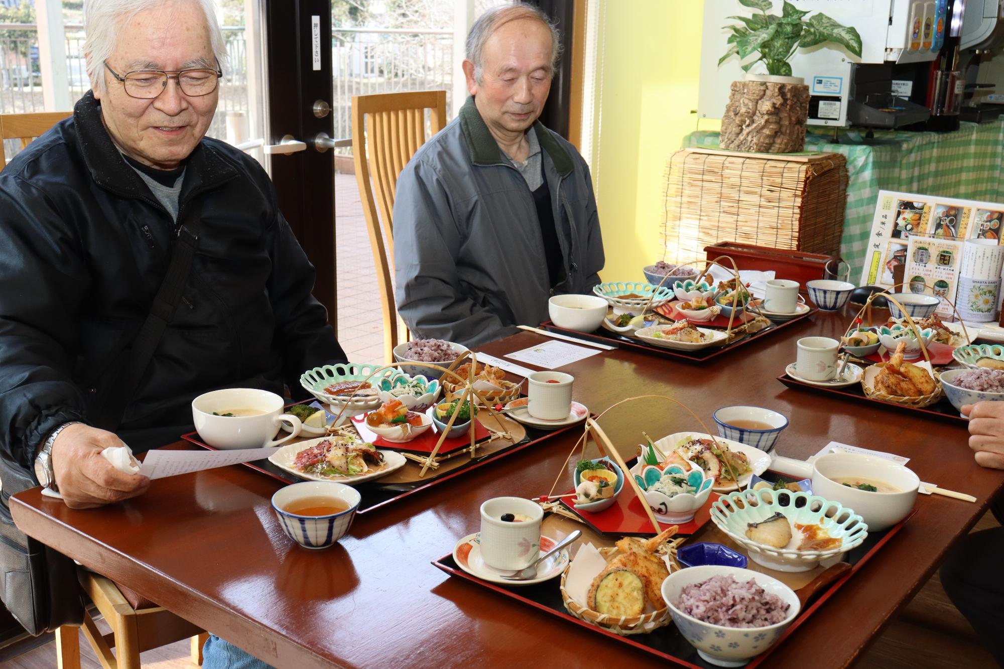 テーブルに運ばれた4人分の伊沢昼御膳を前に小林さんらがお品書きを確認している写真