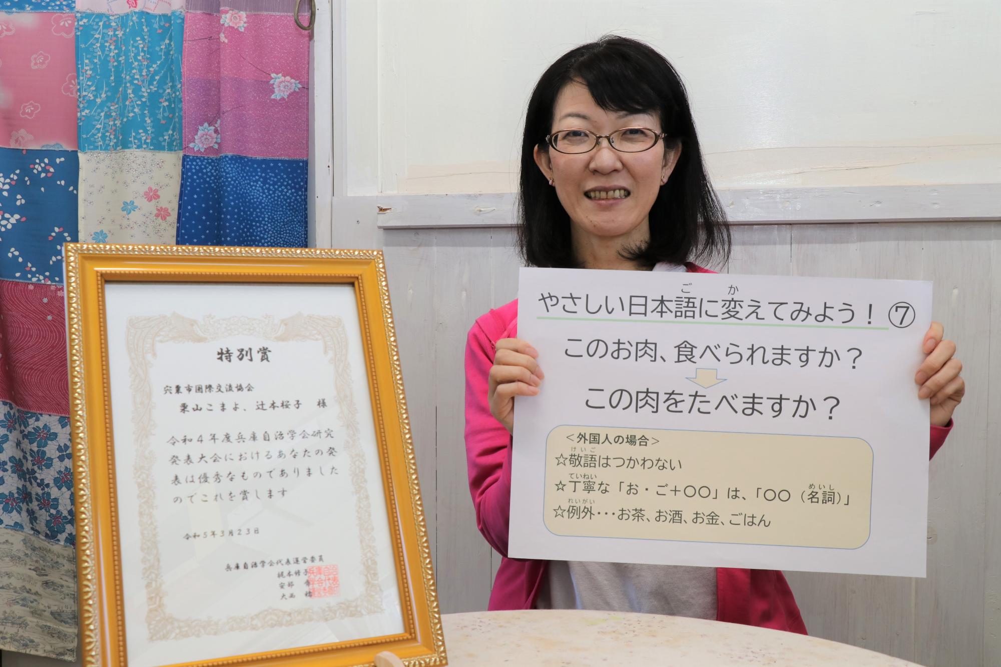 栗山こまよさんが「やさしい日本語」に関する問題が書かれた紙を手に持ち、机の上に立てられた兵庫自治学会特別賞の賞状と一緒に写っている写真