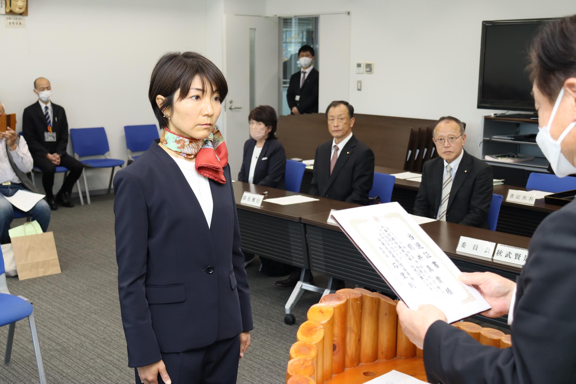 県議選に当選した前井真紀氏（中央）に付与する当選証書を秋武賢是委員長（右）が読み上げている写真