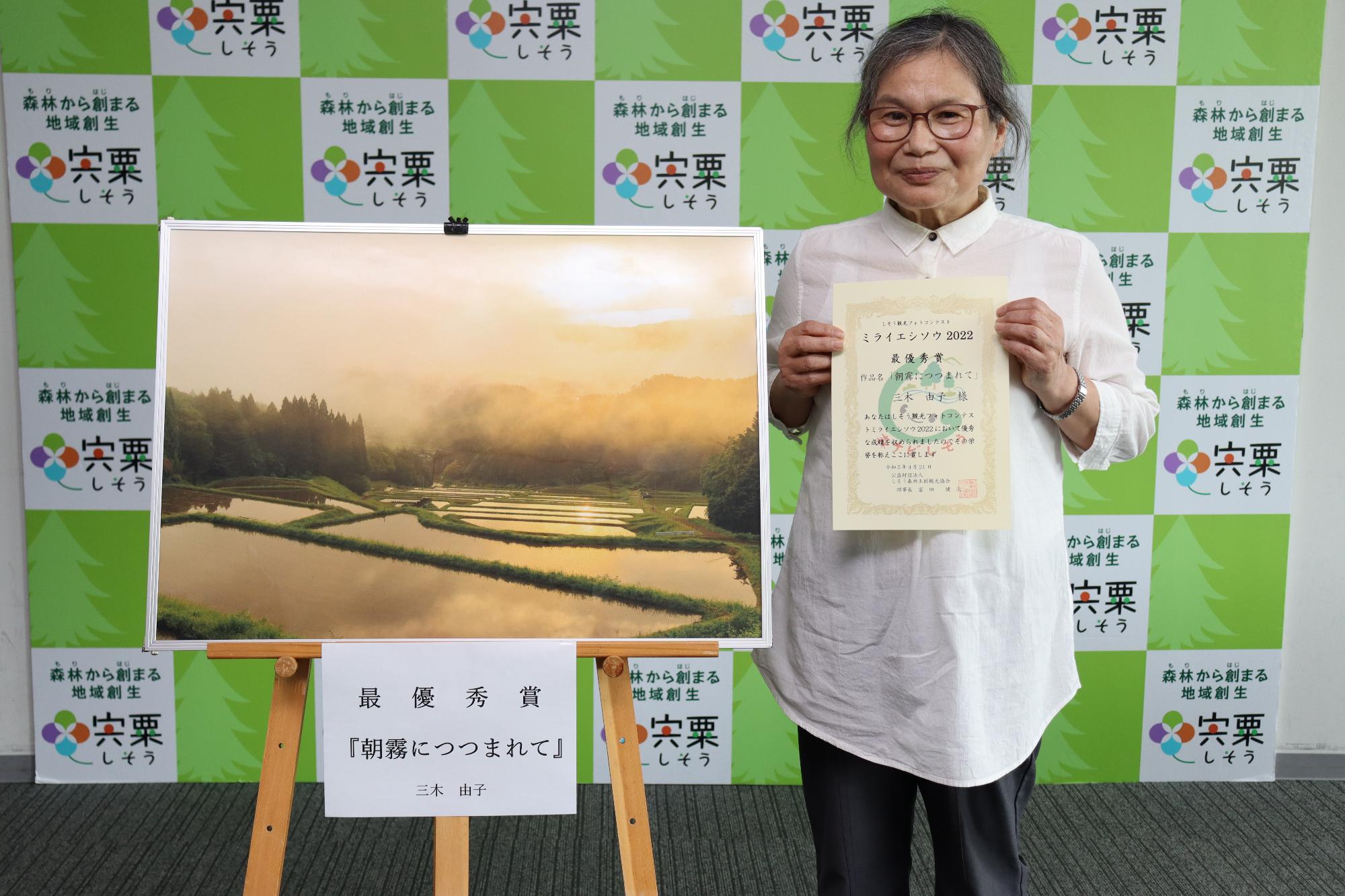 観光協会フォトコンテスト「ミライエシソウ2022」最優秀作品の隣に受賞者の三木さん賞状を手に正面を向いて立っている写真