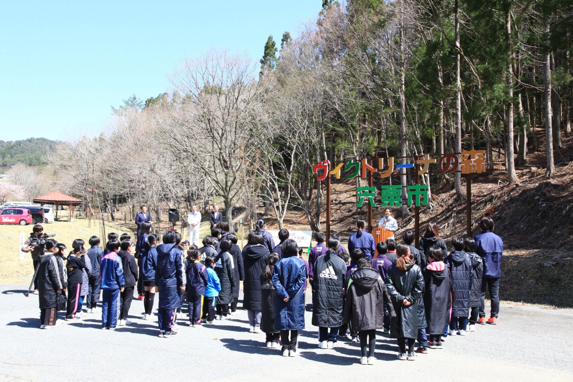 ちくさ高原「ヴィクトリーナの森」看板枚に参加者が集まり福元市長のあいさつを聞いている写真