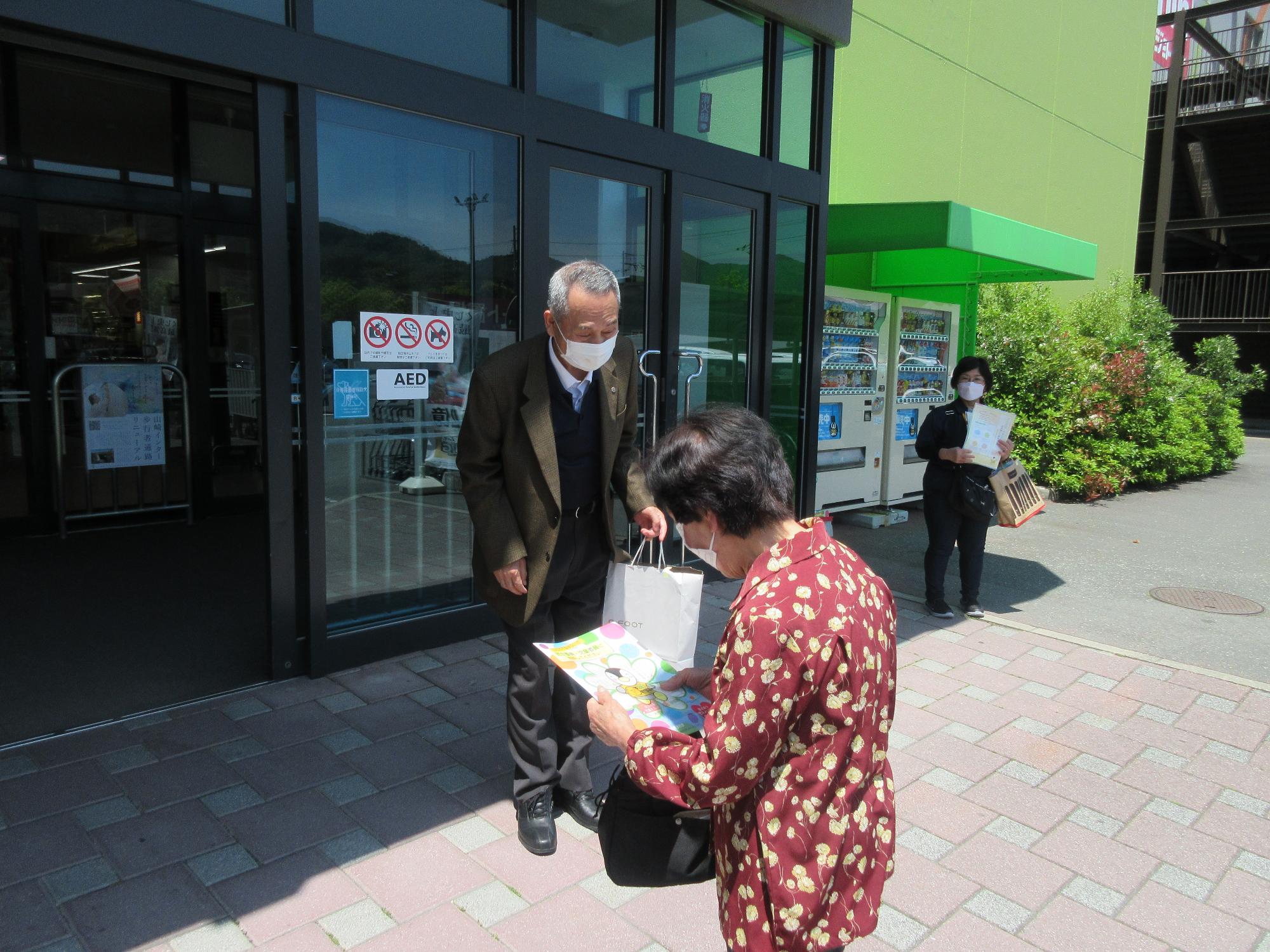 市の民生委員・児童委員がイオン山崎店の店内出入口で配布するチラシを受け取り、目を通している来店者の写真