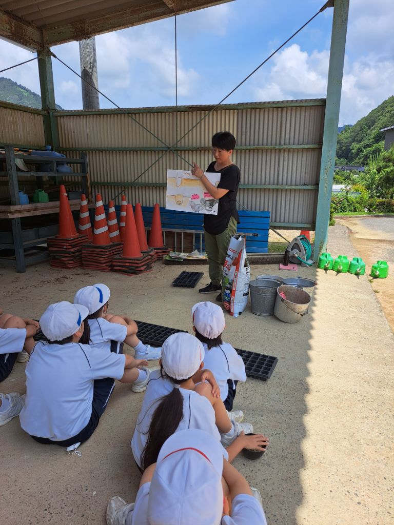蔦沢小学校の3年生が集まり大豆の種のまきかたを指導するスタッフから聞いている写真