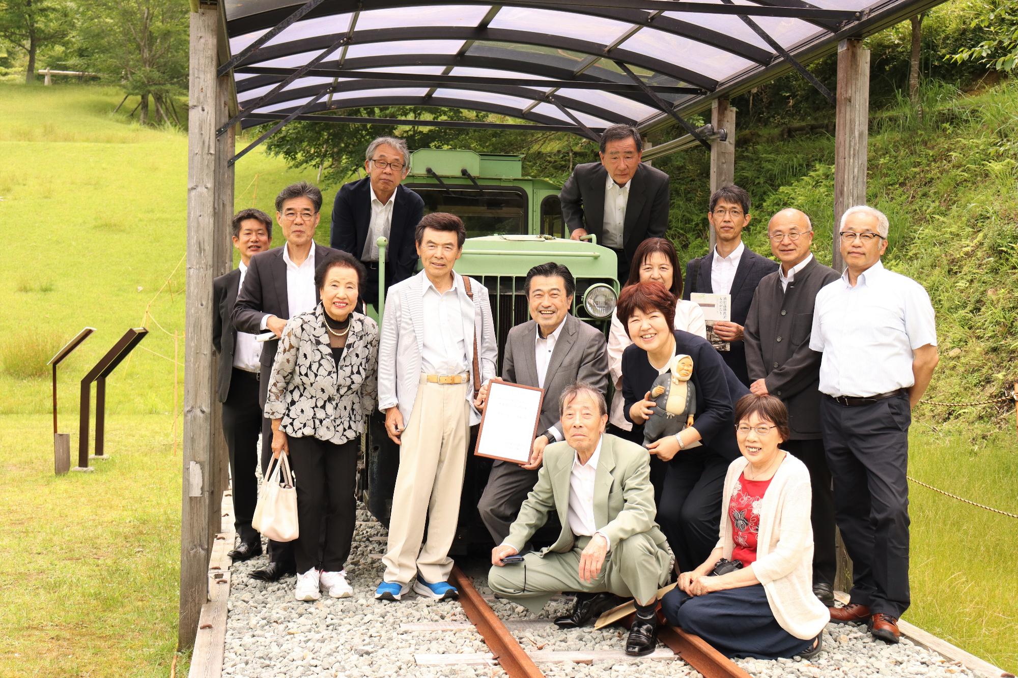 森林鉄道の復活運行をめざす機関車の前で、波賀元気ネットの松本会長が賞状を手に、協議会メンバーや産業遺産学会の小西理事長、鈴木さんとともに記念撮影した写真