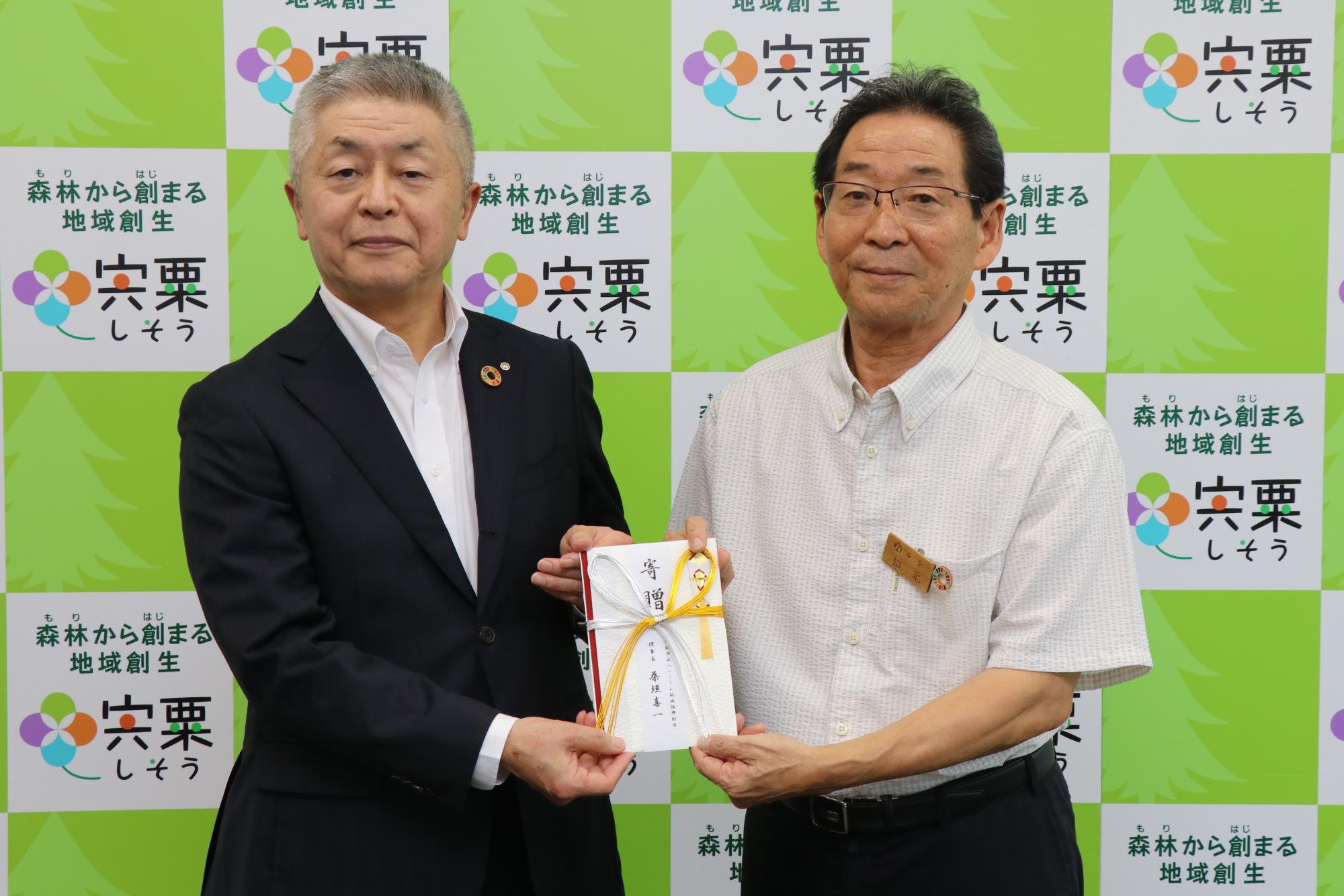 西兵庫信用金庫の桑垣理事長（左）と福元市長が寄贈目録の四角を持ち、正面に掲げて前を向いている写真