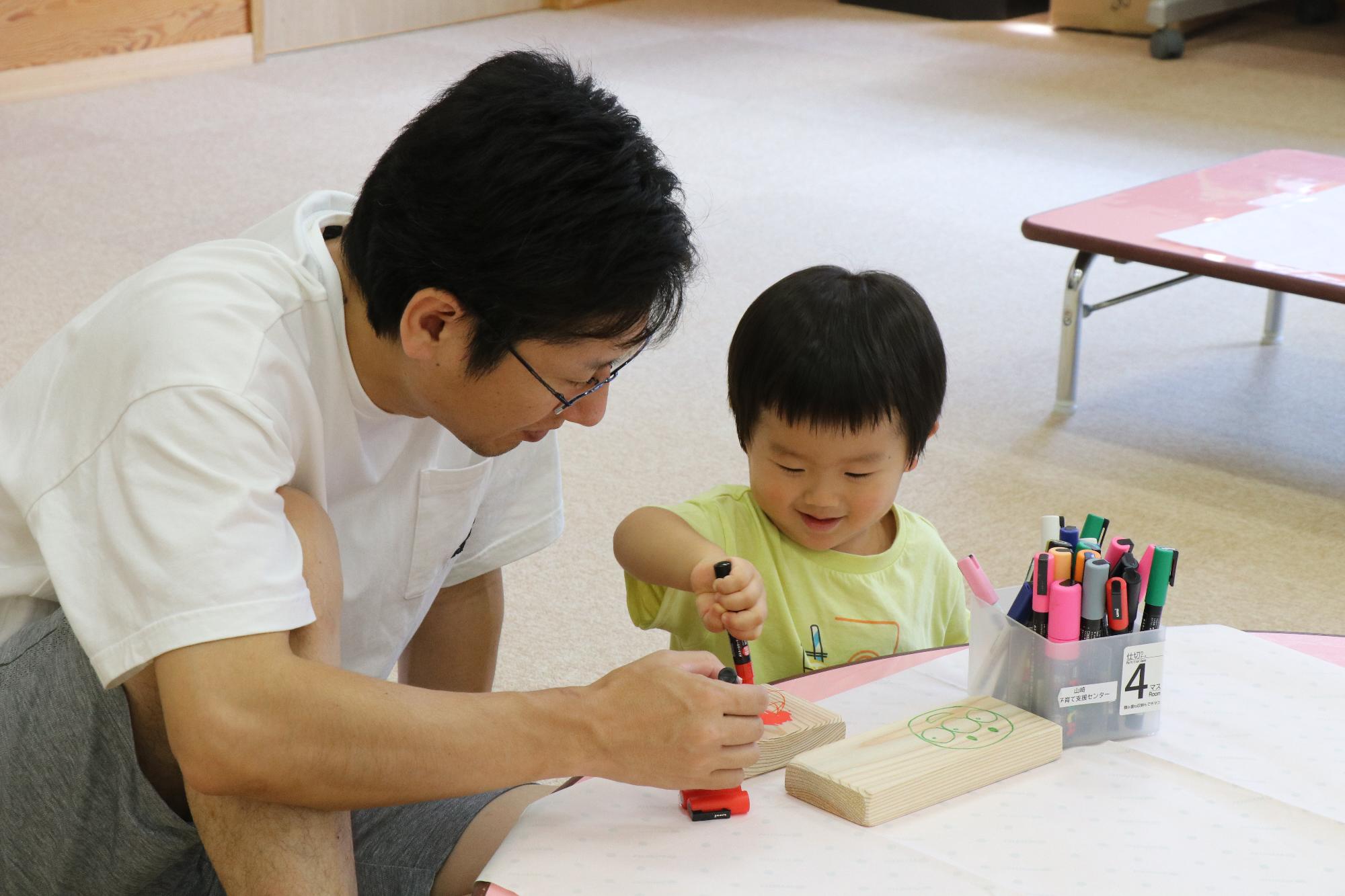 親子が座って小さな木の板にマジックで一緒に絵を描いている写真