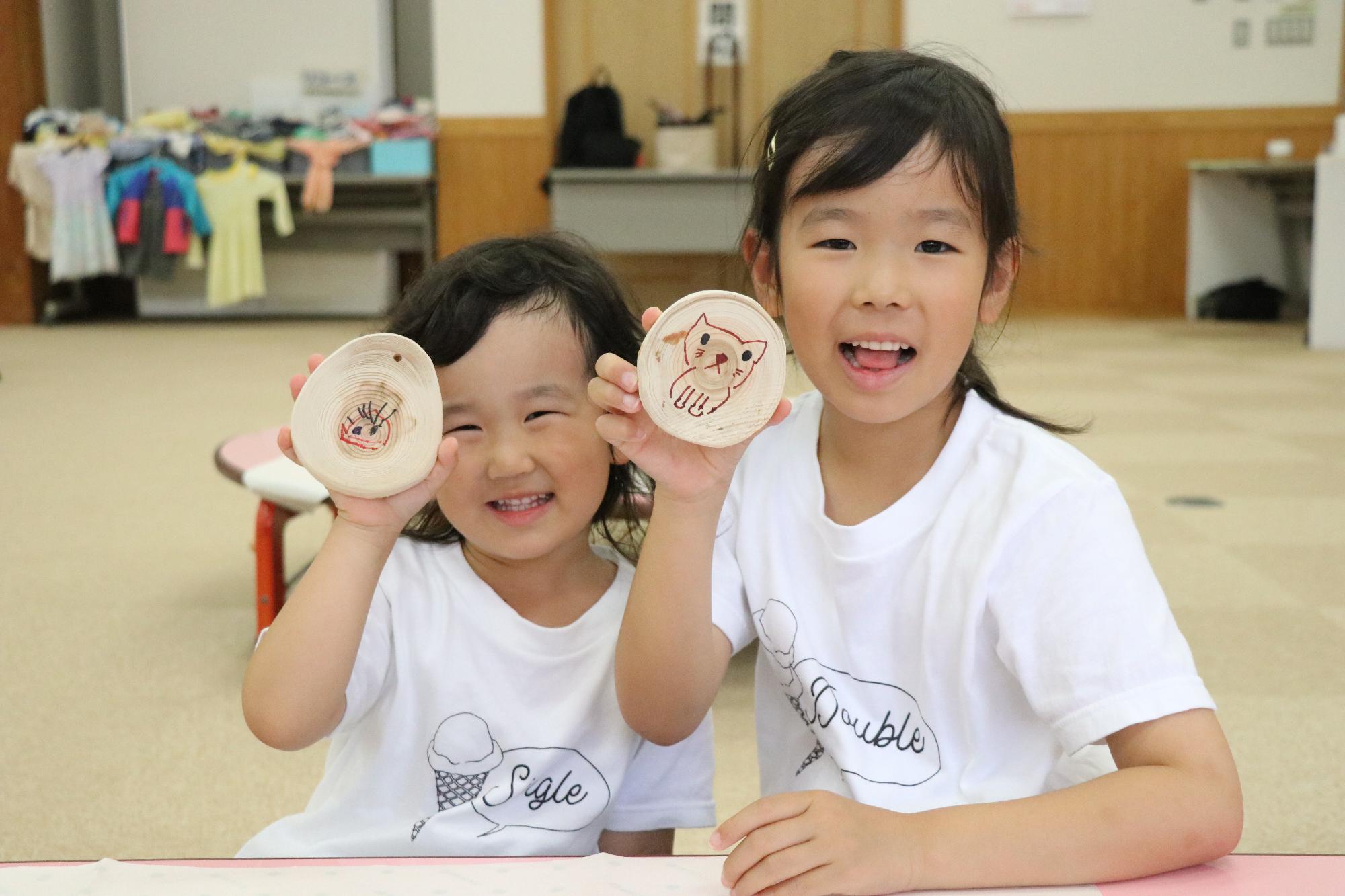 子ども二人が並んで座って自分たちが木の板に描いた絵を笑顔で見せている写真