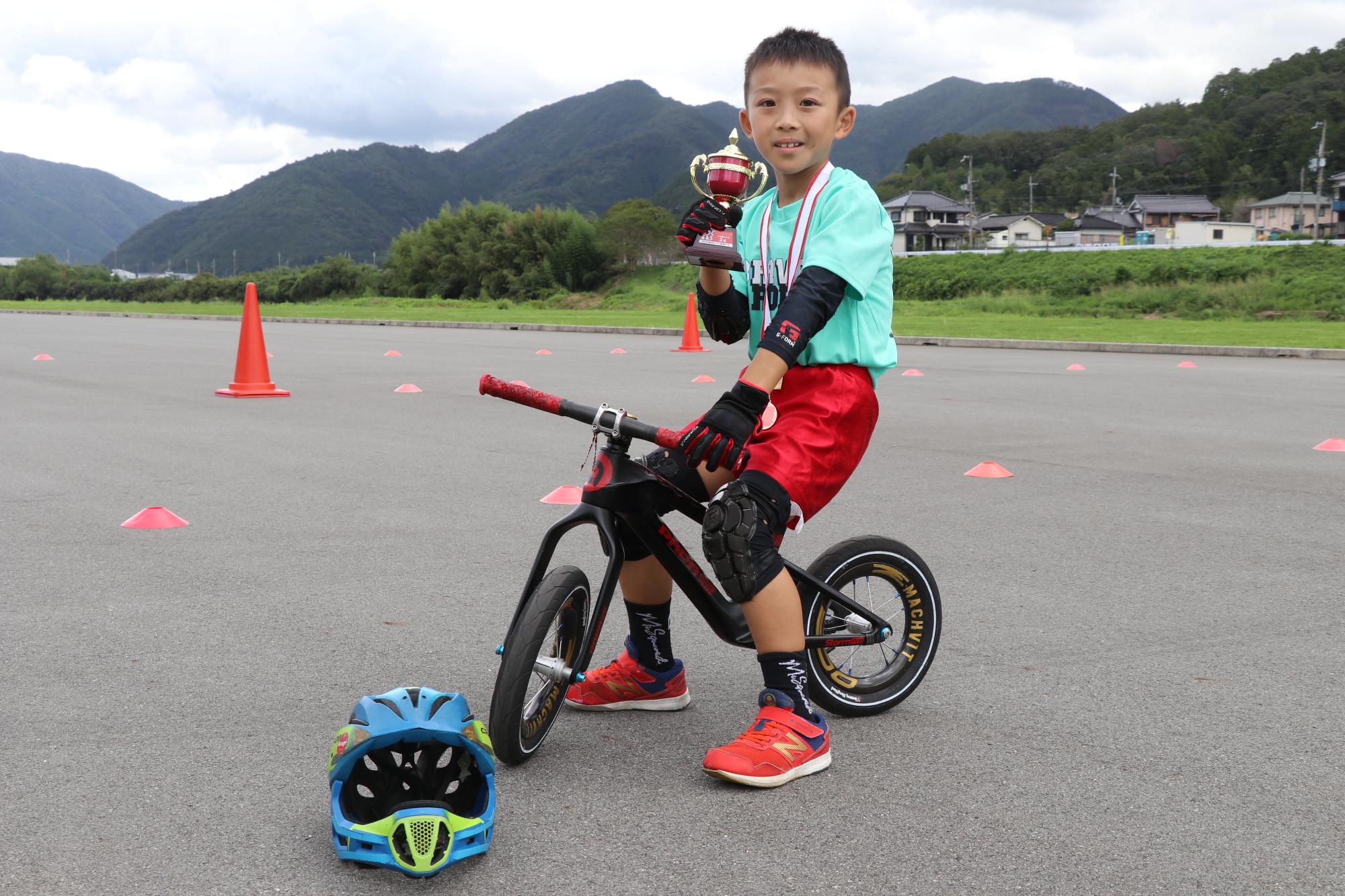 せせらぎ公園で植田さんがランバイクにまたがり、3位入賞記念のカップを持っている写真