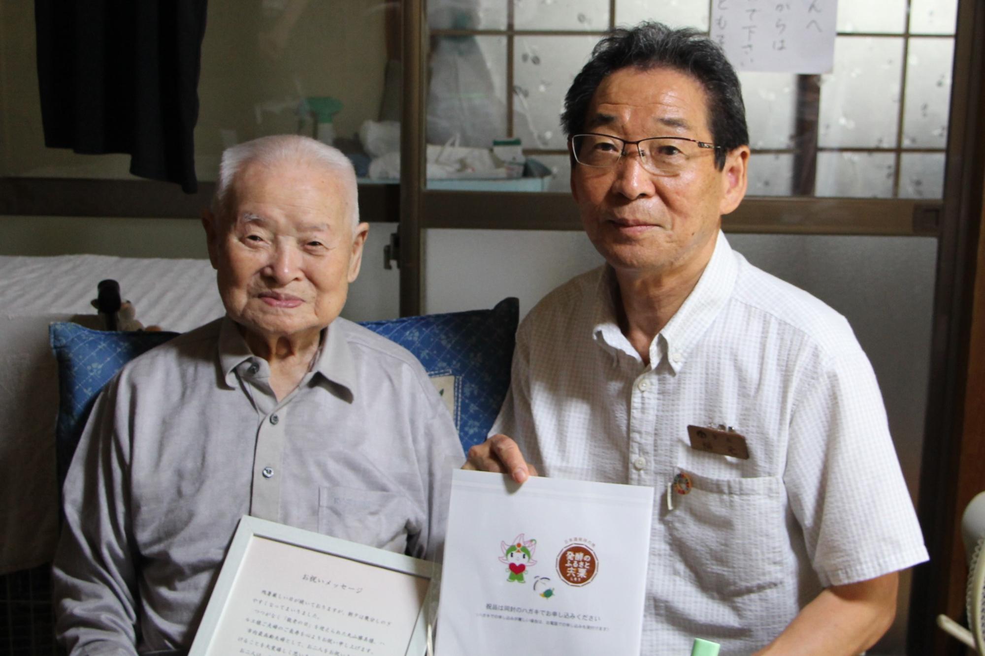 最高齢夫婦の夫である丸山勝美さんが市長から受け取ったお祝いのメッセージを手に市長と並んで座っている写真