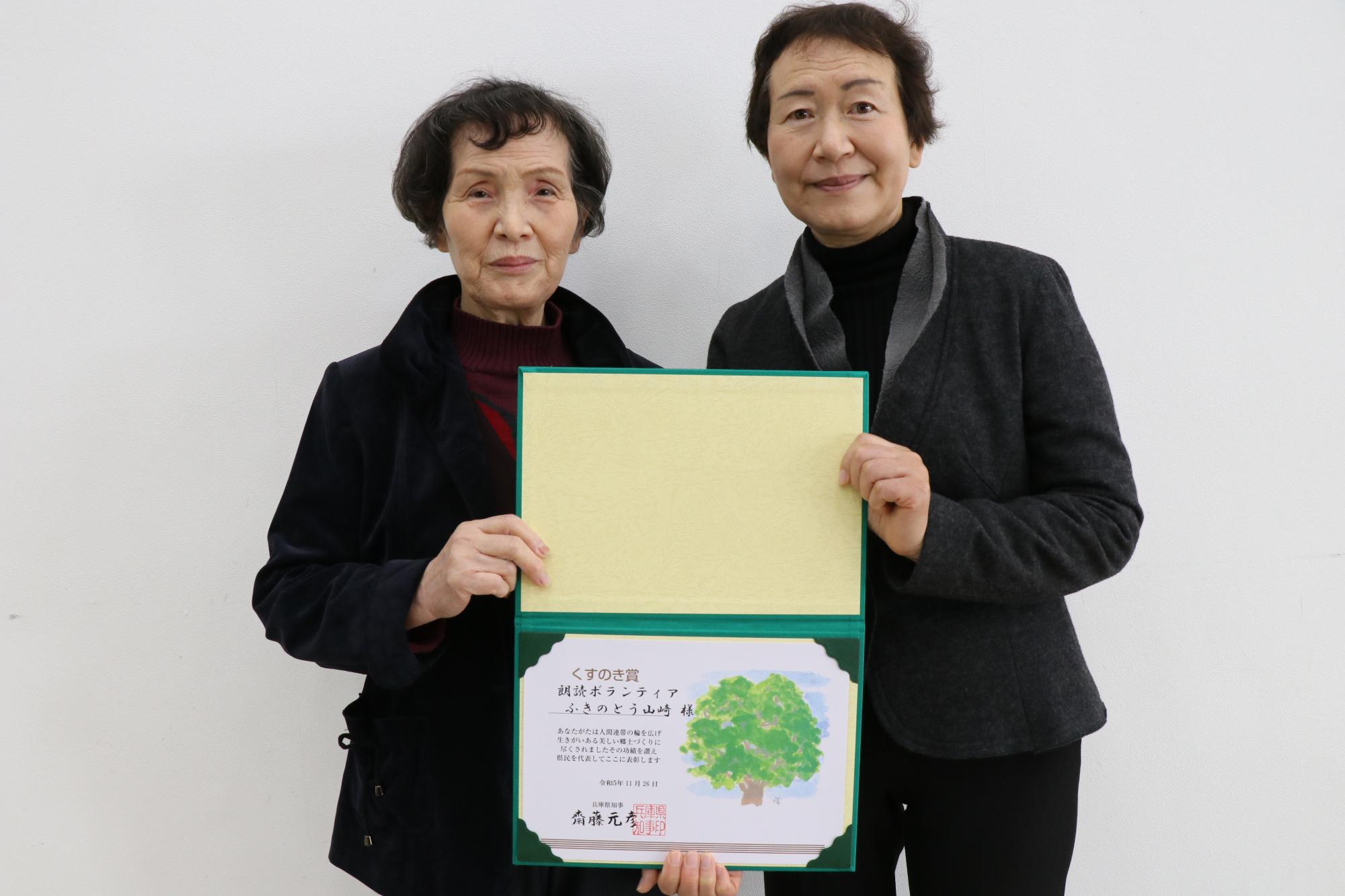 朗読ボランティアふきのとう山崎の古河さん（左）と同団体の前野さんが一緒にくすのき賞の賞状を手に持ち笑顔をみせている画像
