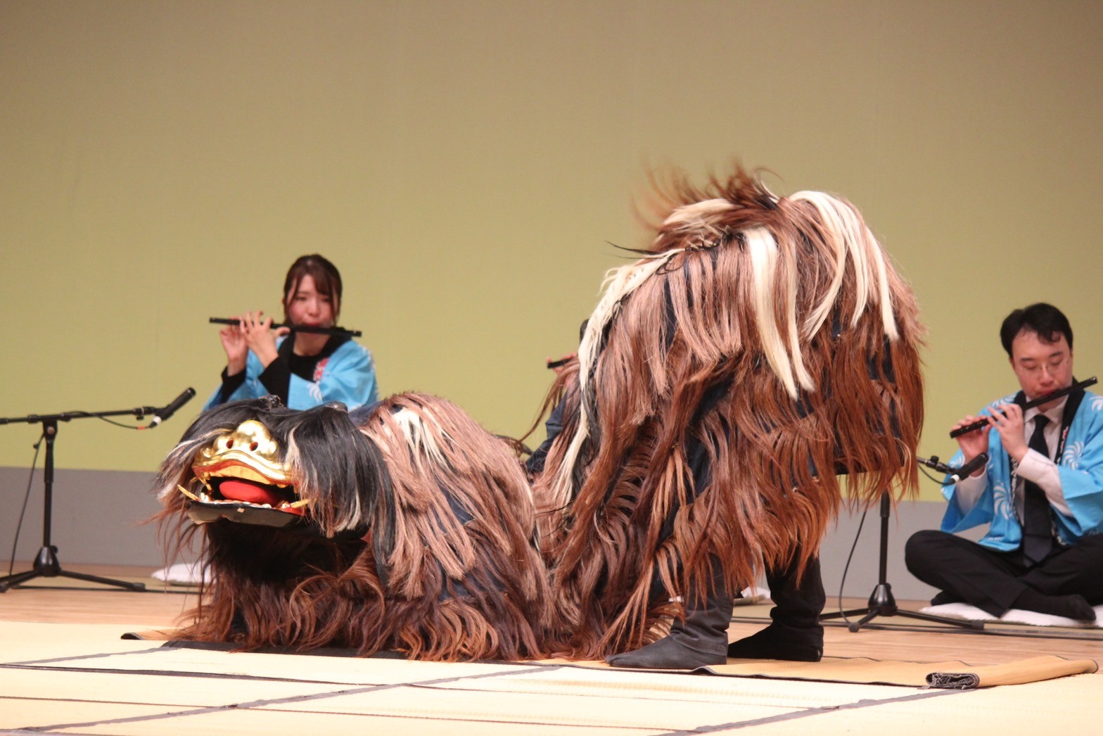 丸尾建築あすかホールの舞台で、宇原獅子舞保存会が獅子舞と篠笛を披露している写真