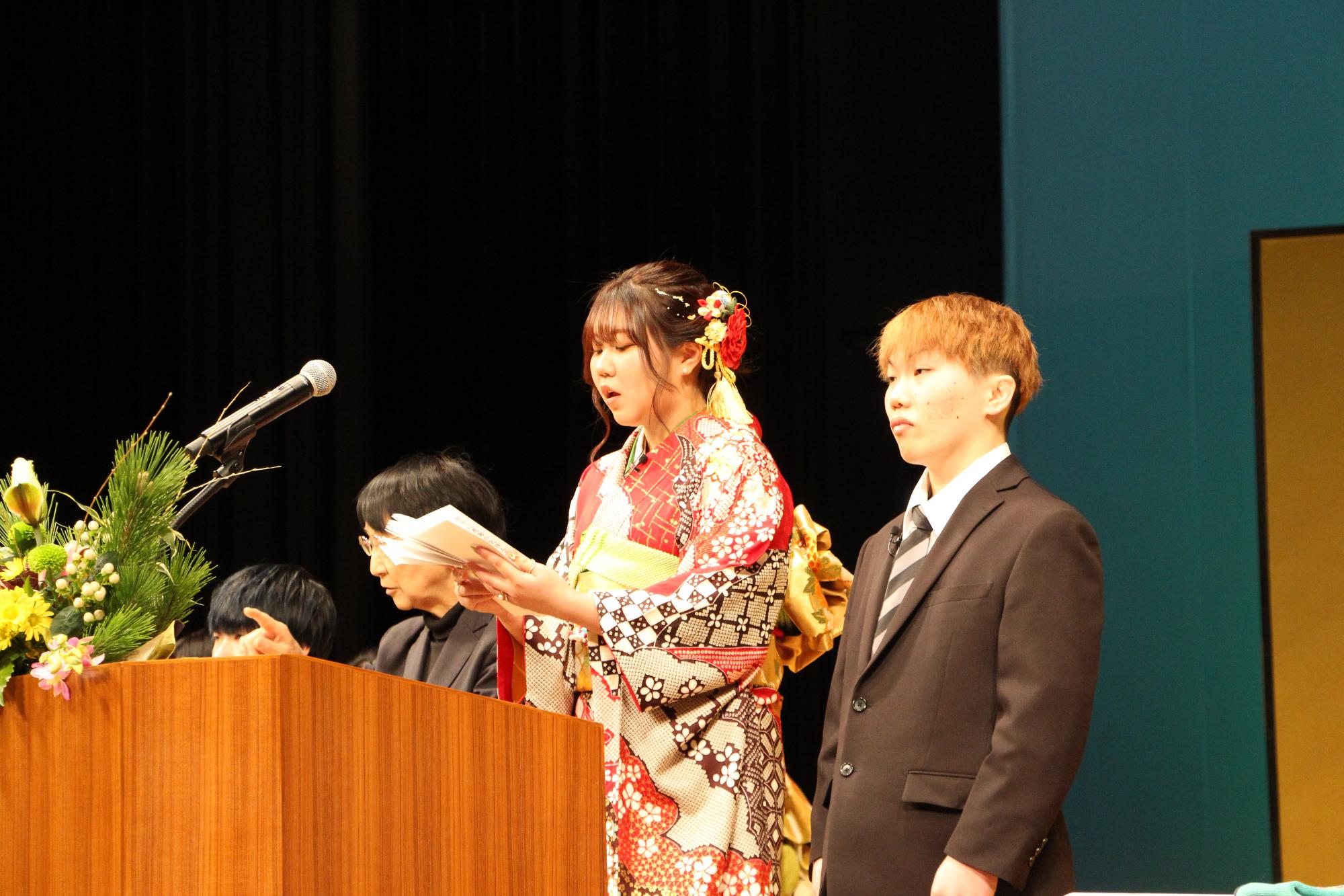 山崎文化会館のステージ上で新成人らに向かって誓いの言葉を述べる多賀成美さんと西村亘貴さんの写真