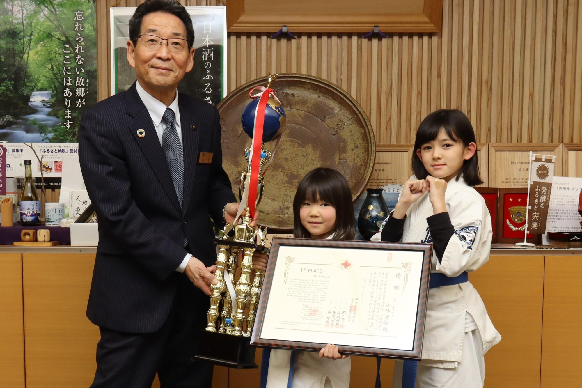 優勝トロフィーと賞状を手に持った小林さんを真ん中に、坂井さんと福元市長が隣に並んで記念撮影した写真