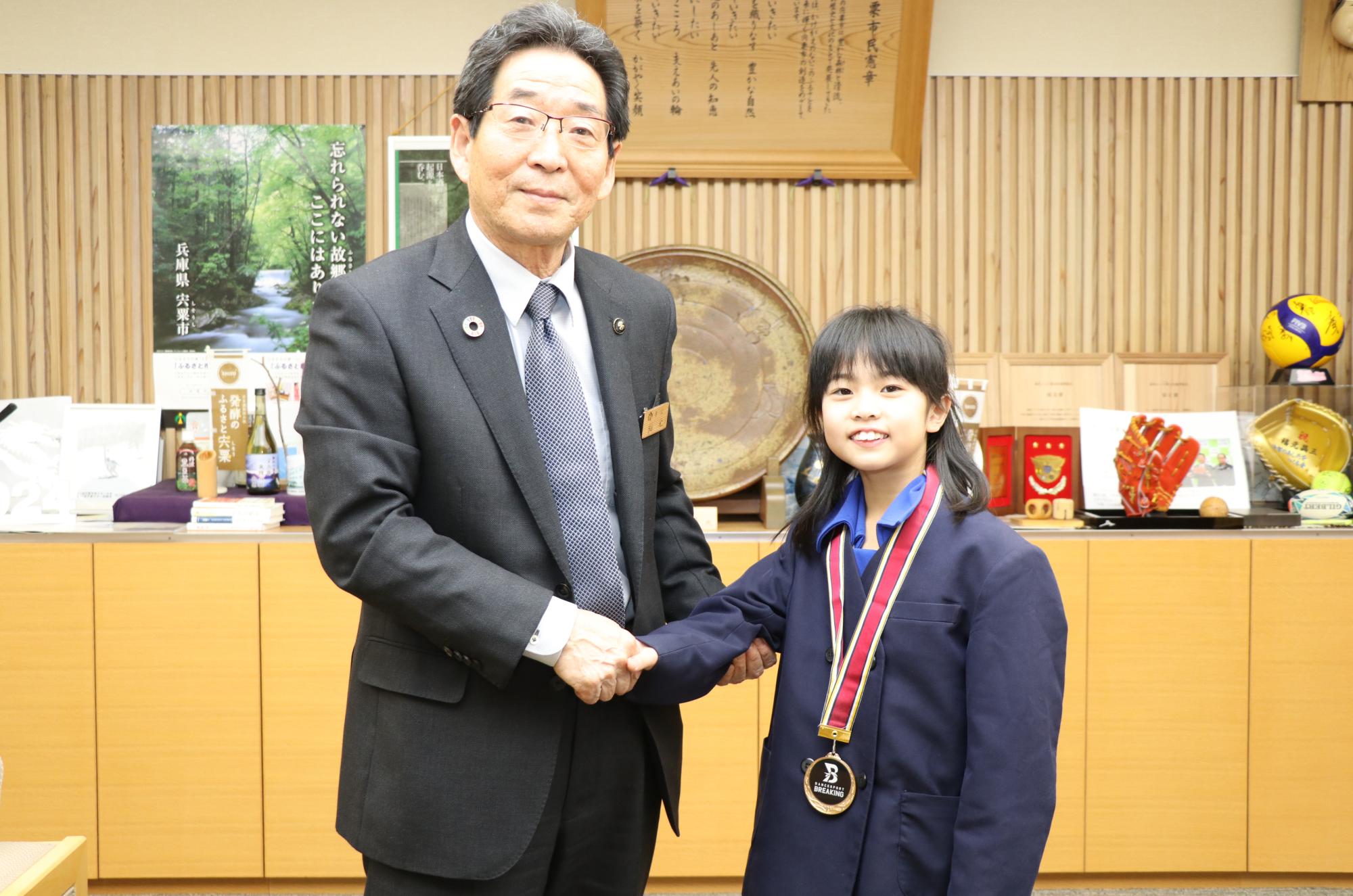 優勝メダルを首にかけた宇田さんと福元市長が市長室で横に並び、握手している写真