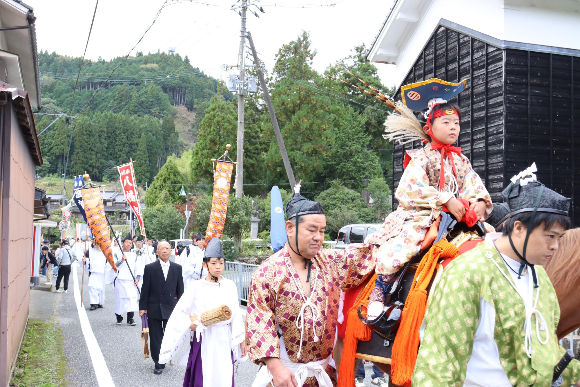 馬に乗った男児に続いて祭礼道具を持った人たちが列をなして道を歩いている写真