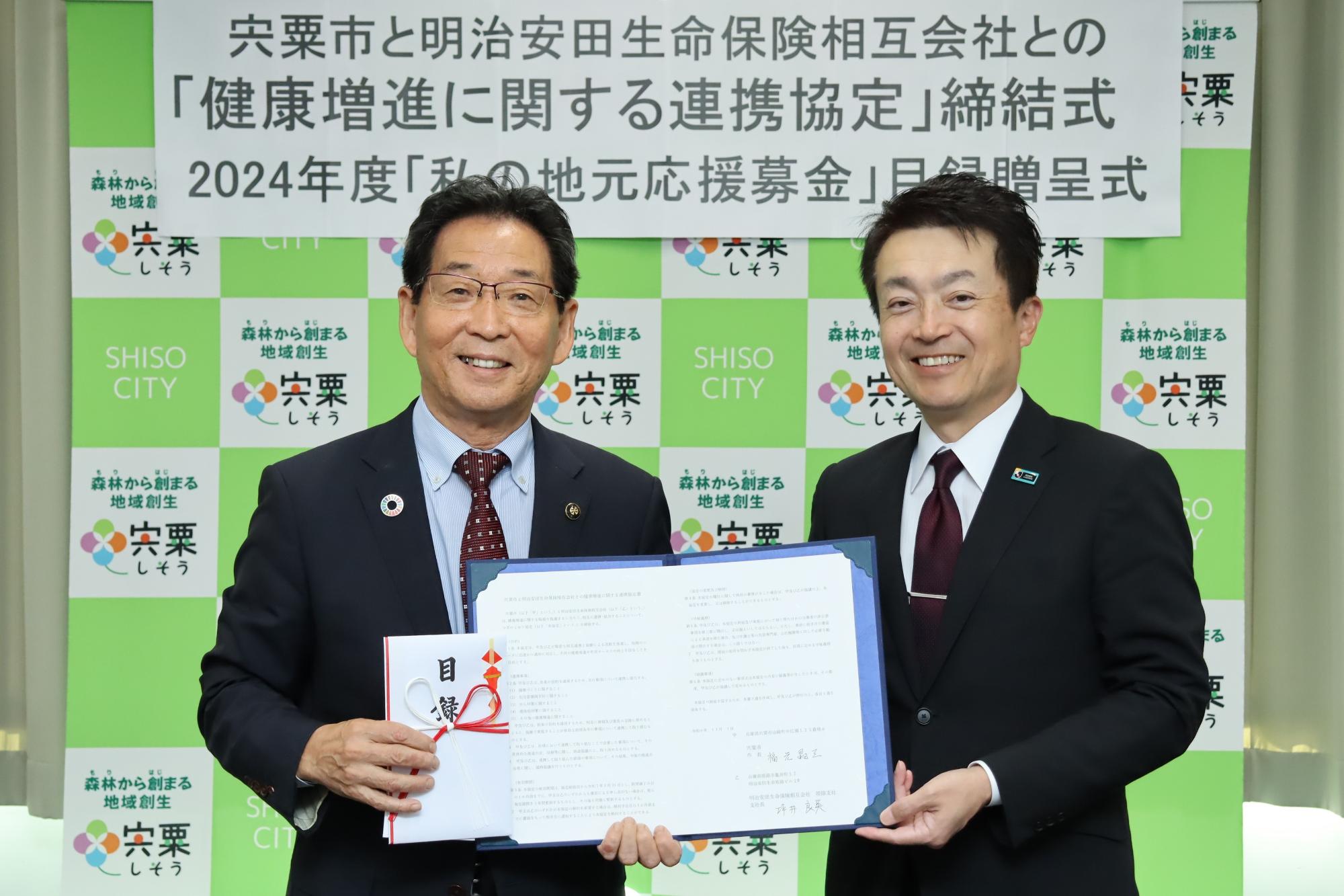 明治安田生命保険相互会社の坪井良英姫路支社長と福元市長が取り交わした協定書と目録を掲げて正面を向いている写真