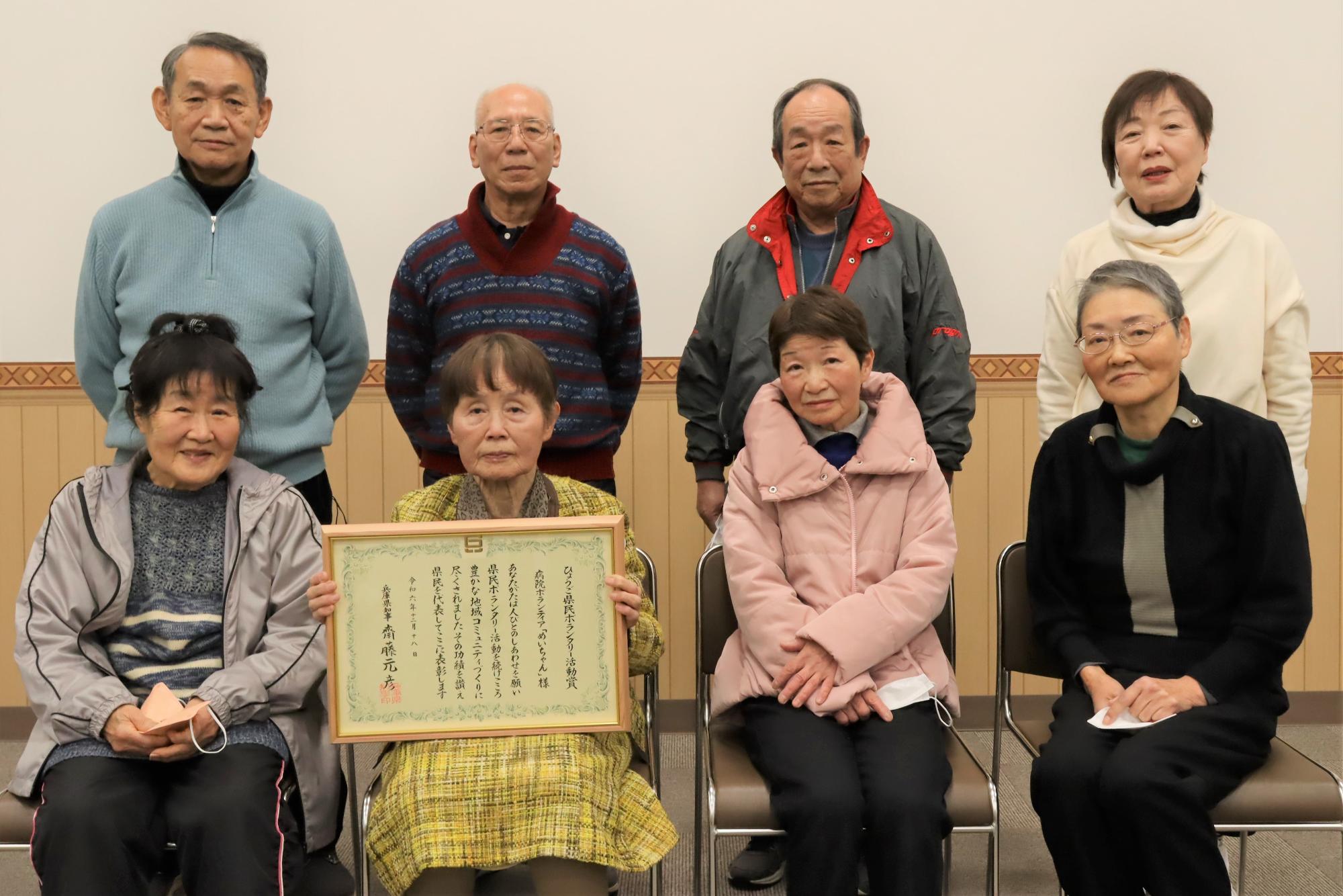 病院ボランティア「めいちゃん」メンバーの集合写真で、イスに4人が座り、ほかの4人が立った状態で前後2列に並んで正面を向いている写真