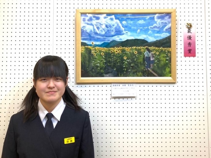 最優秀賞を受賞した作品「向日葵」が額に入って飾られ、作品を描いた田路さんがその前に立って正面を向いている写真